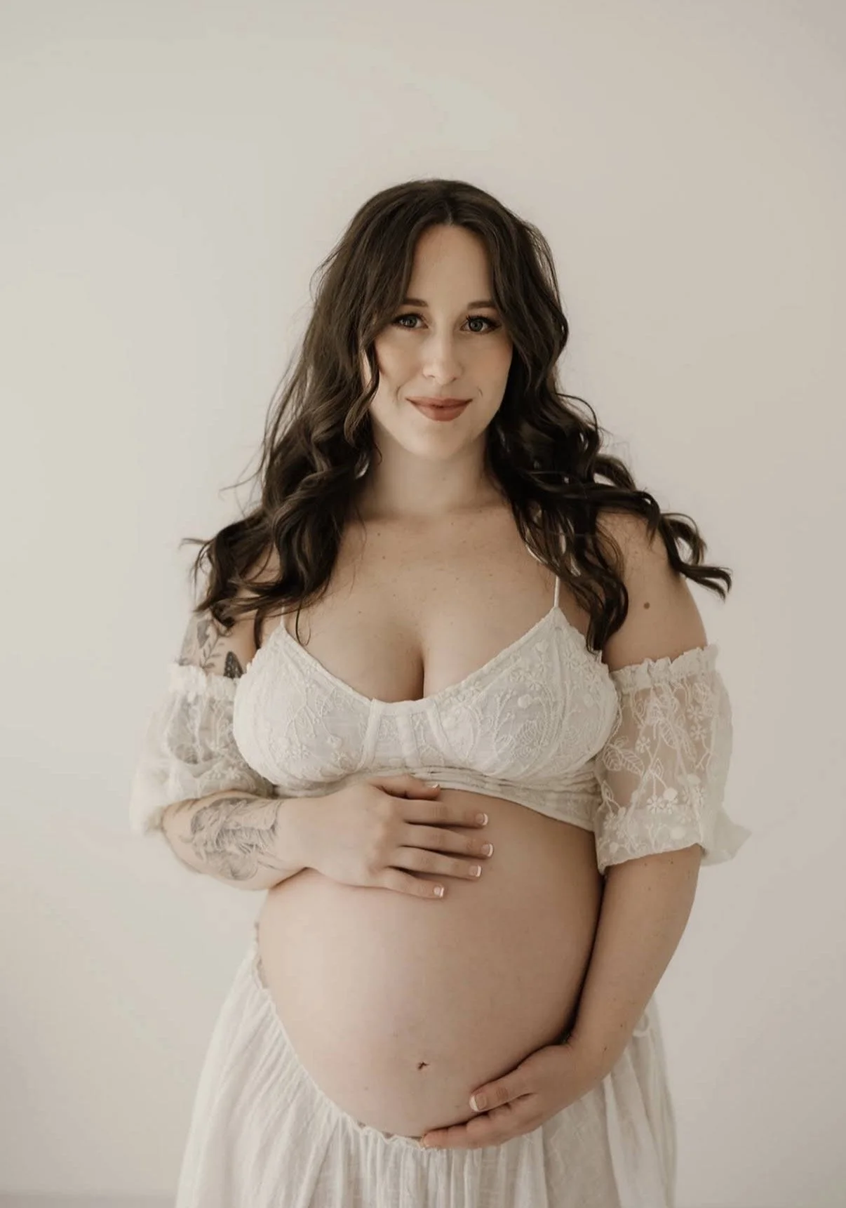 Pregnant woman with long curly brown hair, wearing a white lace bra and matching off-shoulder blouse, standing against a plain light background, gently touching her belly with a soft expression.