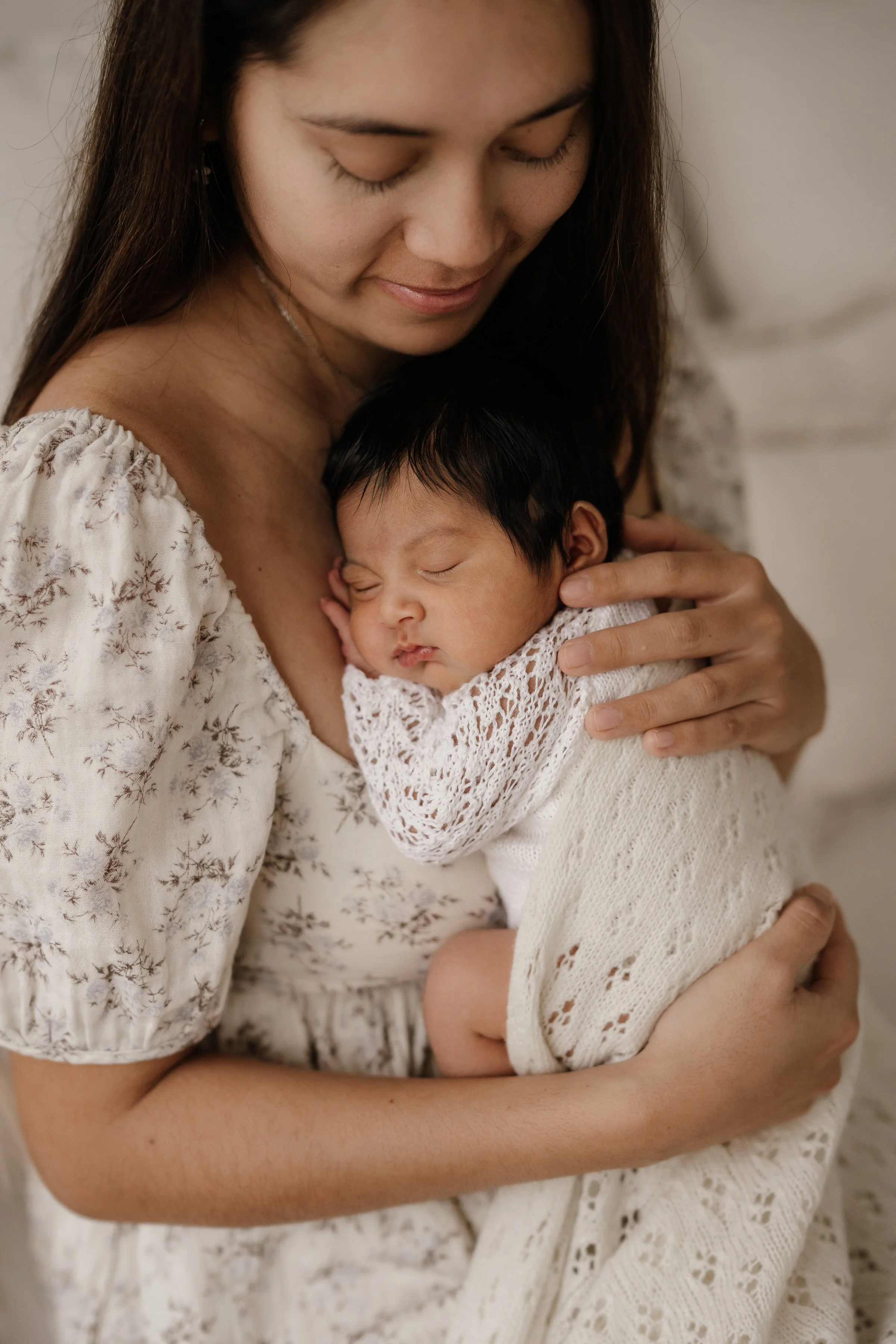 A woman tenderly holding a sleeping baby wrapped in a white, knitted blanket, both wearing matching white outfits with lace and floral patterns.