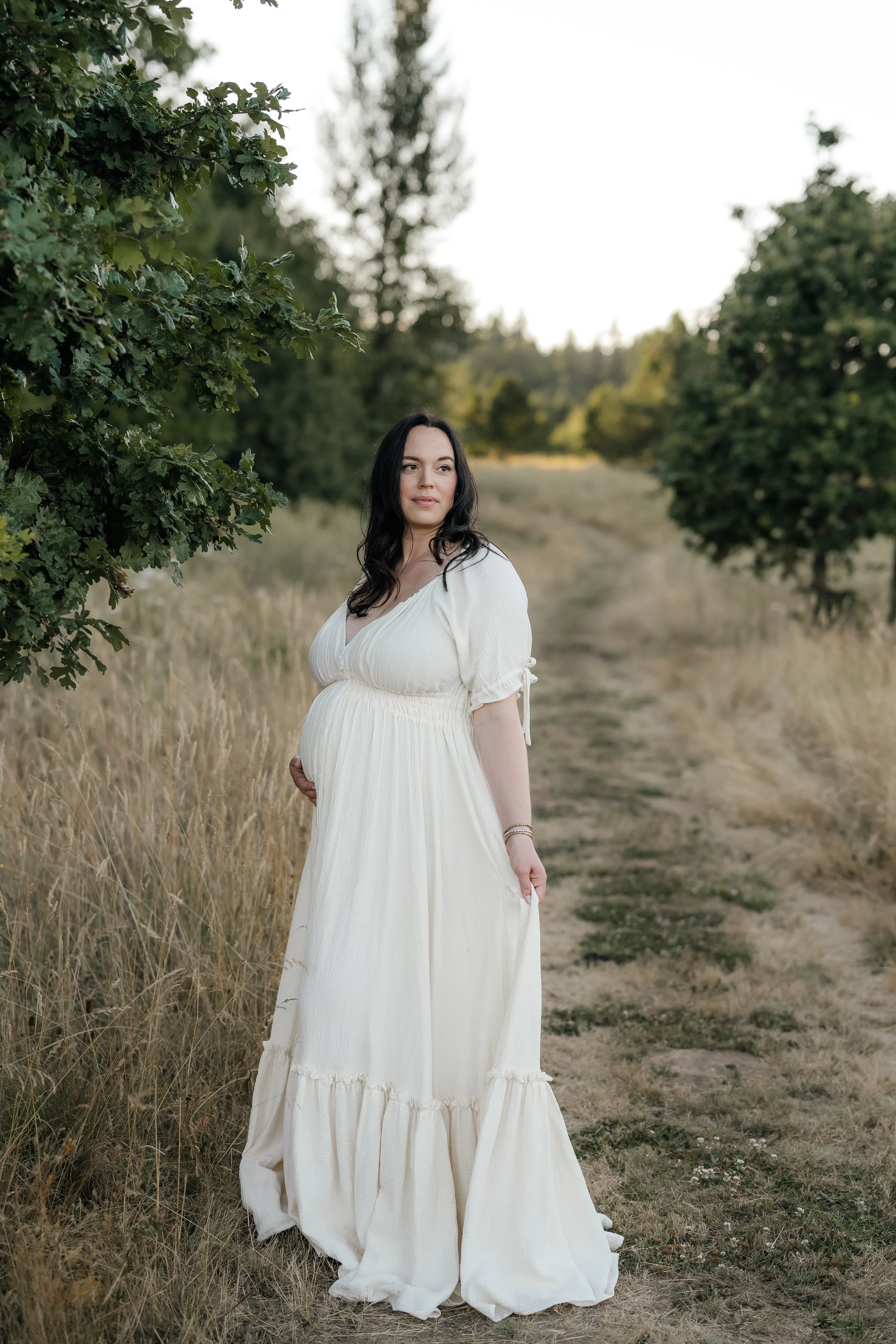 A pregnant woman with long dark hair standing on a dirt path in a grassy field, surrounded by trees, wearing a flowing cream-colored dress.