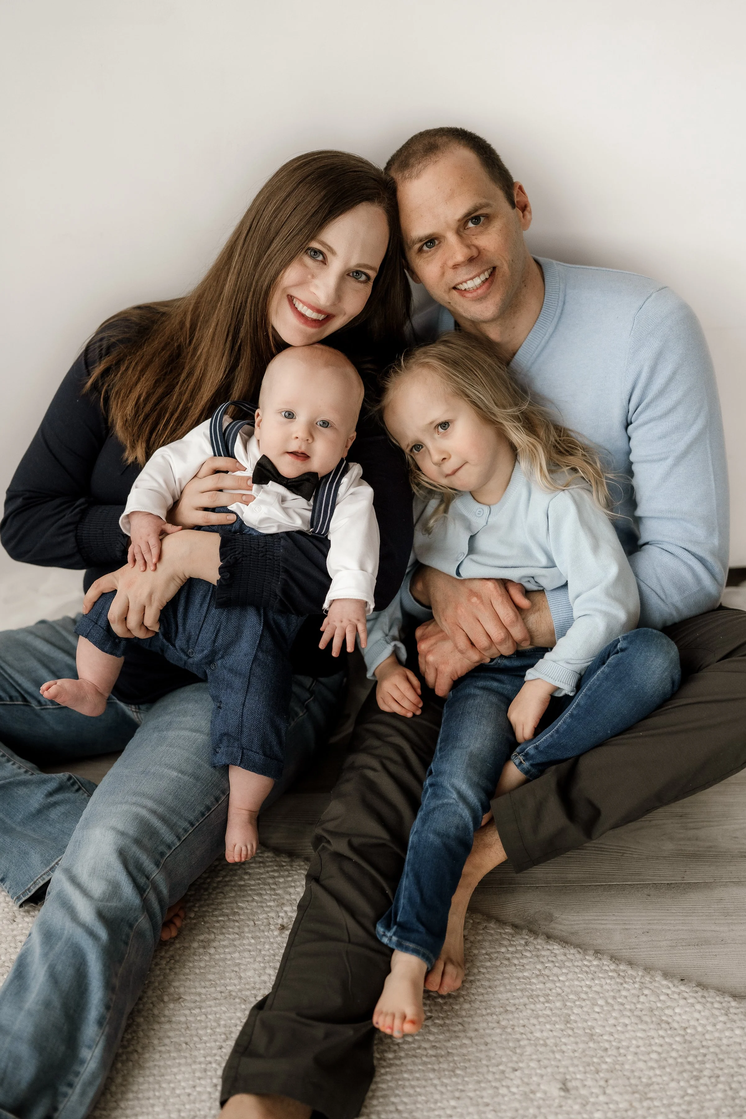 A family of four sitting on the floor in front of a plain white wall. The mother has long brown hair and is smiling, holding their baby boy dressed in a tuxedo with a bow tie. The father has short brown hair and is smiling, with a young girl with lon