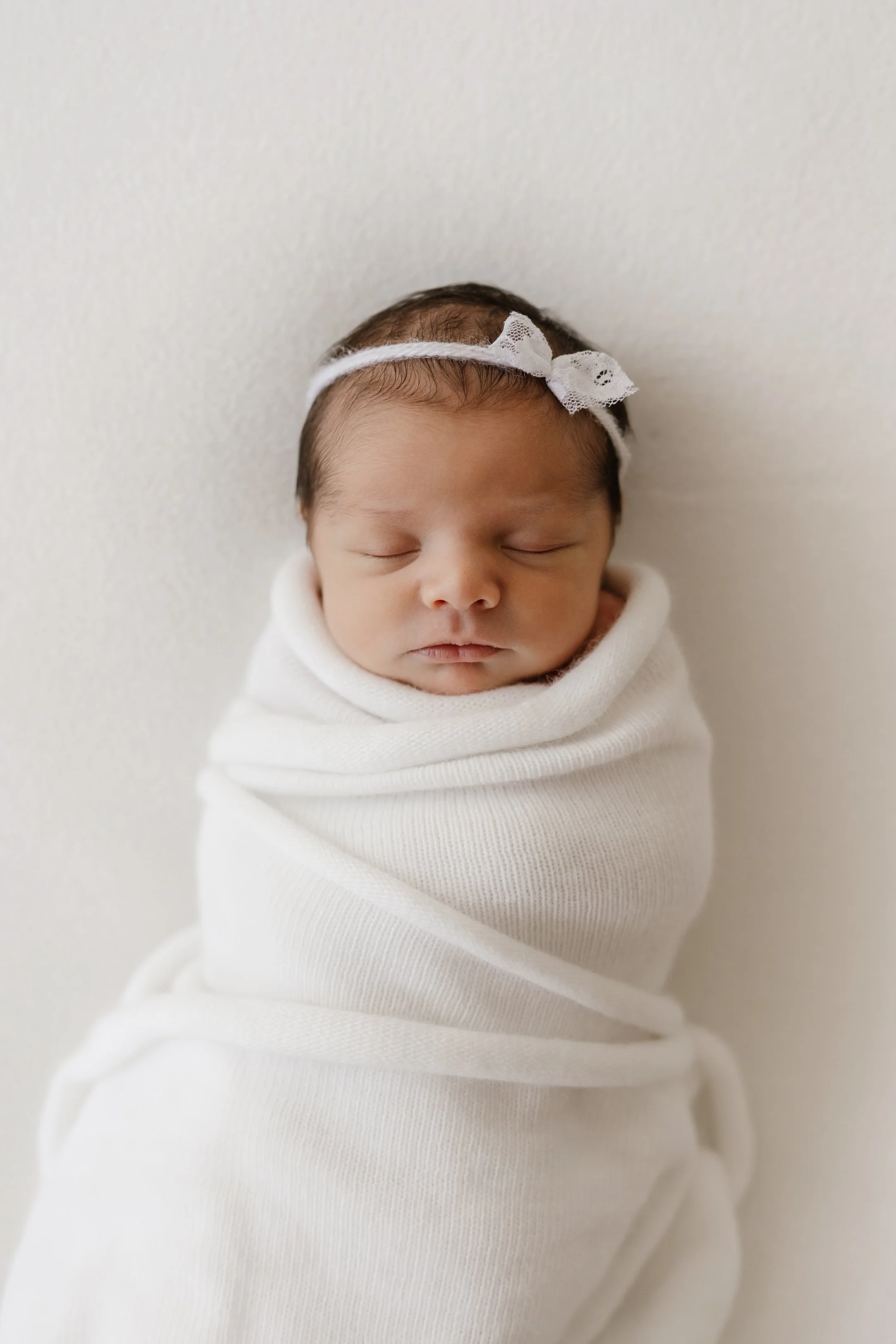 Newborn baby wrapped in a white blanket, sleeping peacefully on a white background, with a white lace headband.