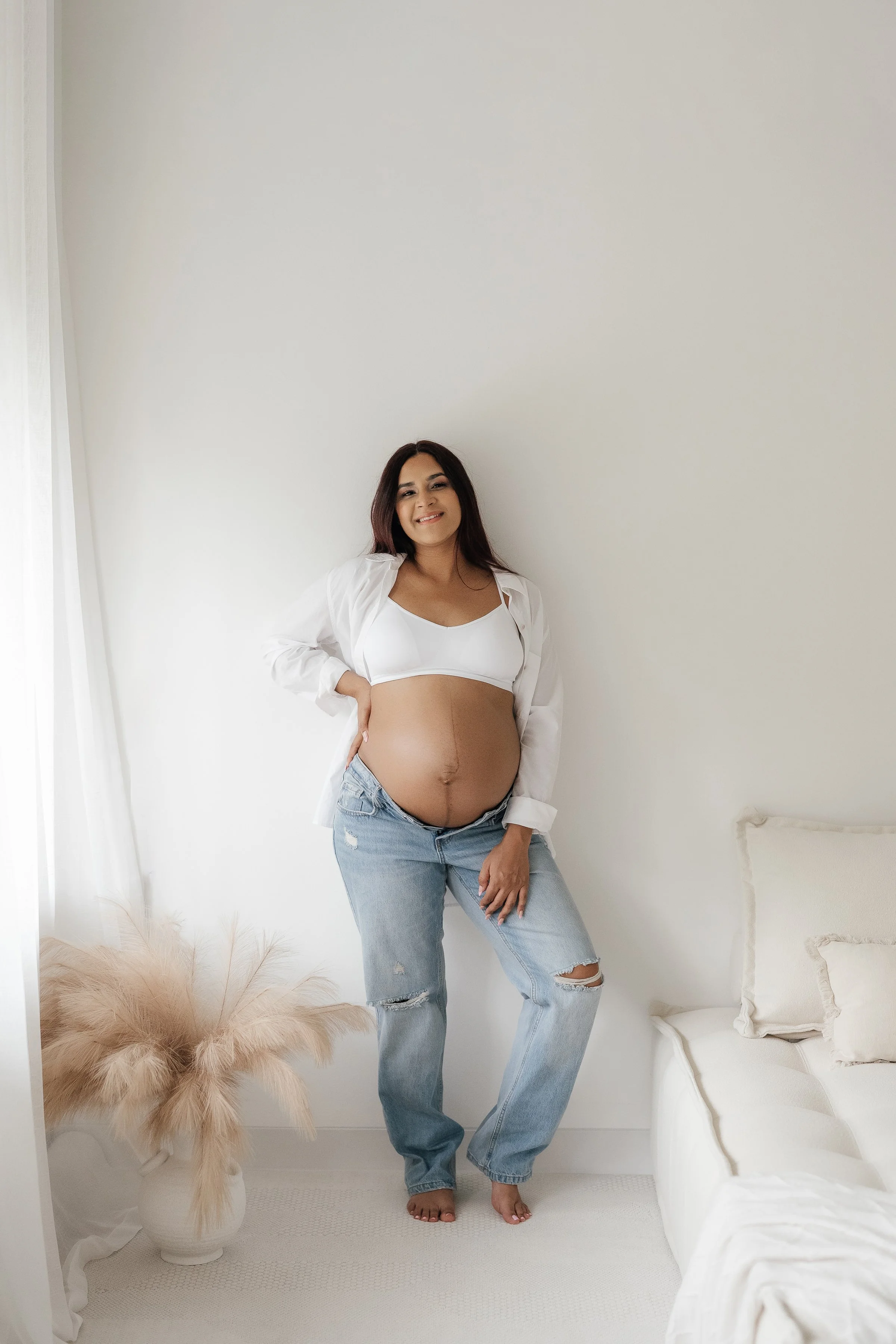 A pregnant woman with long dark hair, wearing a white crop top, oversized white shirt, and ripped jeans, standing barefoot in a bright minimalistic room with white walls, next to a potted pampas grass plant and a white sofa.