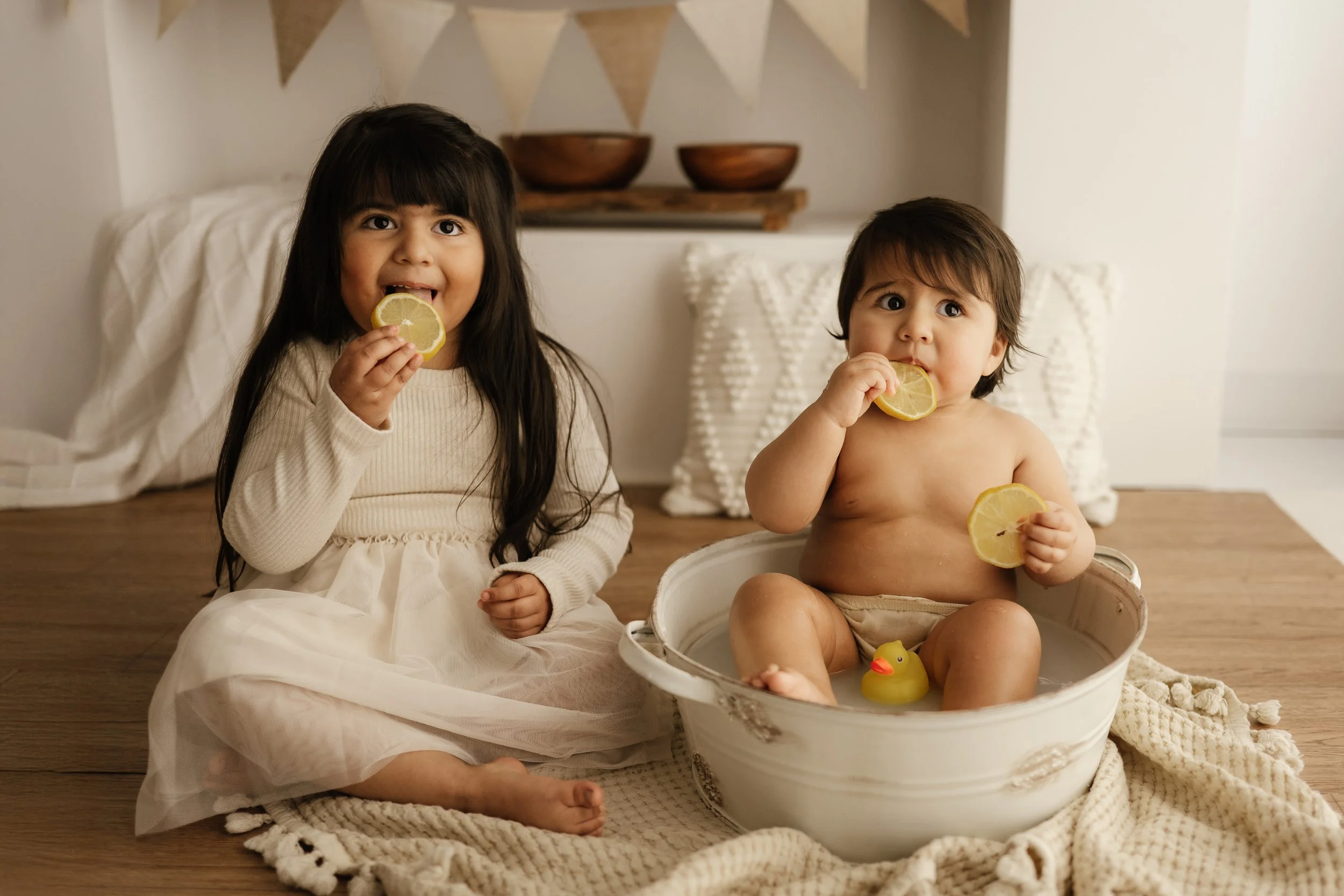 Two young children, a girl and a toddler boy, sitting on a wooden floor with a white blanket. They are eating lemon slices and the boy is sitting in a white basin. The girl is wearing a cream dress, and the boy is shirtless with a rubber duck nearby.
