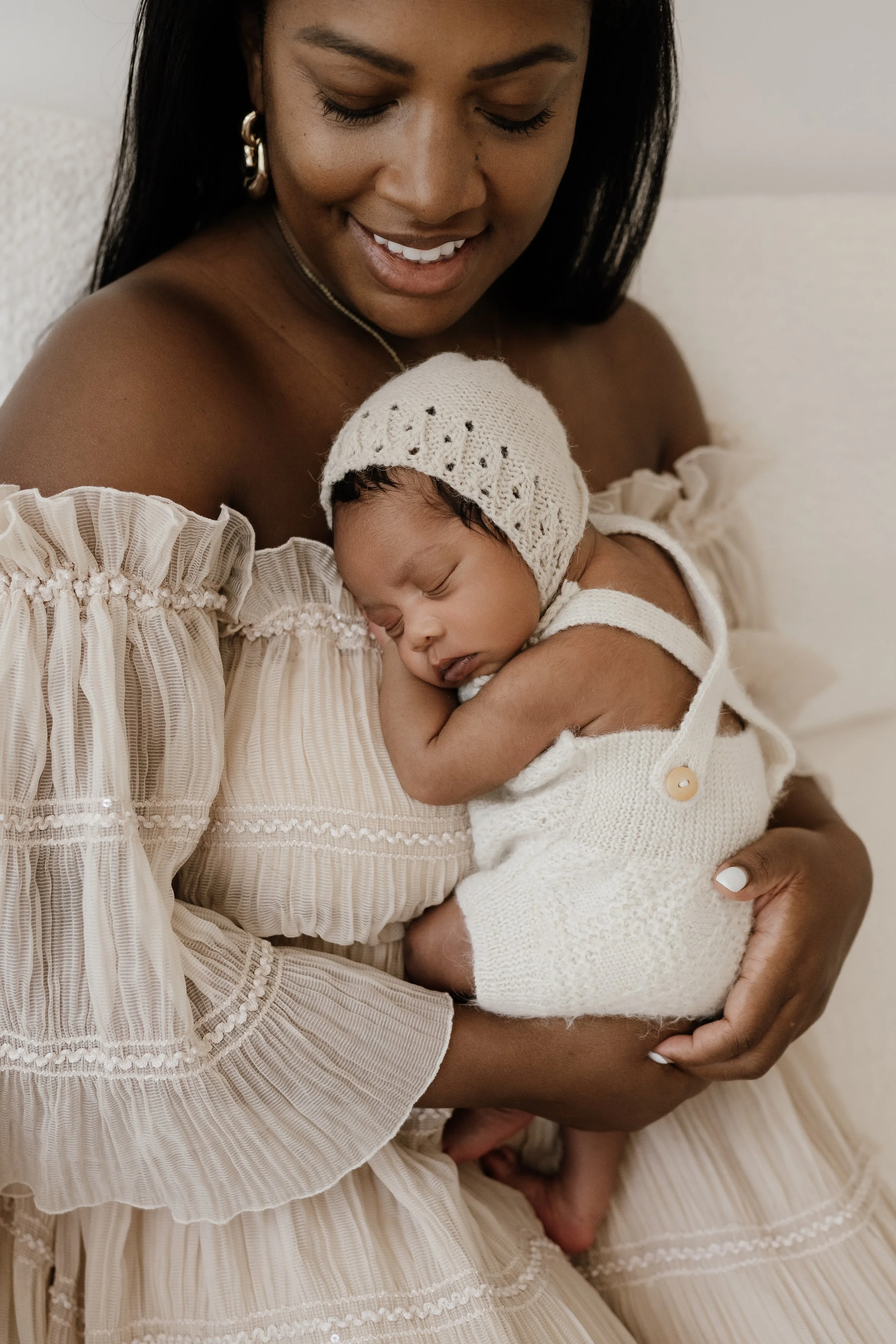 A woman in a beige, ruffled dress holding a sleeping baby in a knitted outfit and hat, smiling gently.