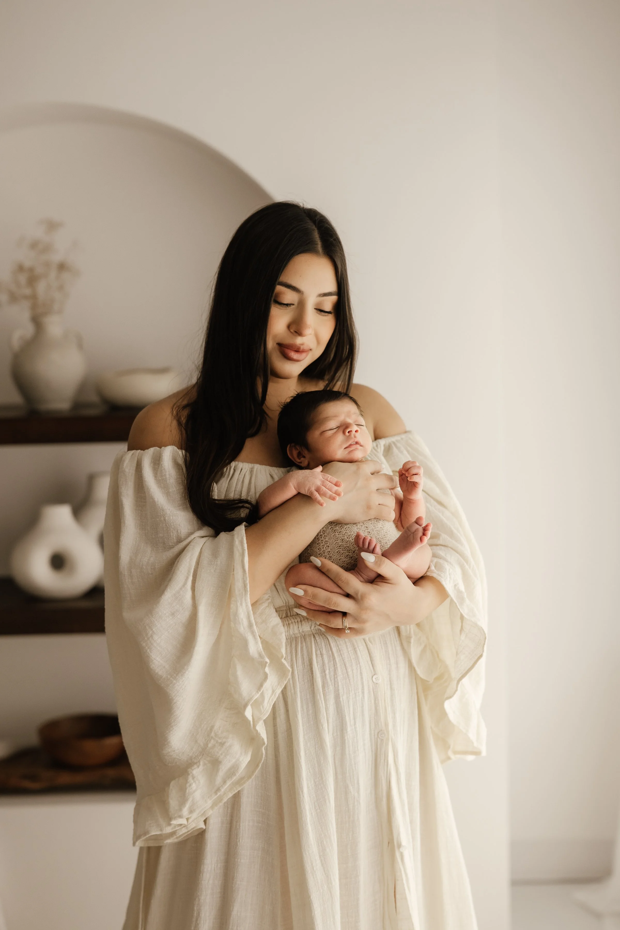 A woman holding a newborn baby in a well-lit, minimalistic room with decorative vases on shelves.