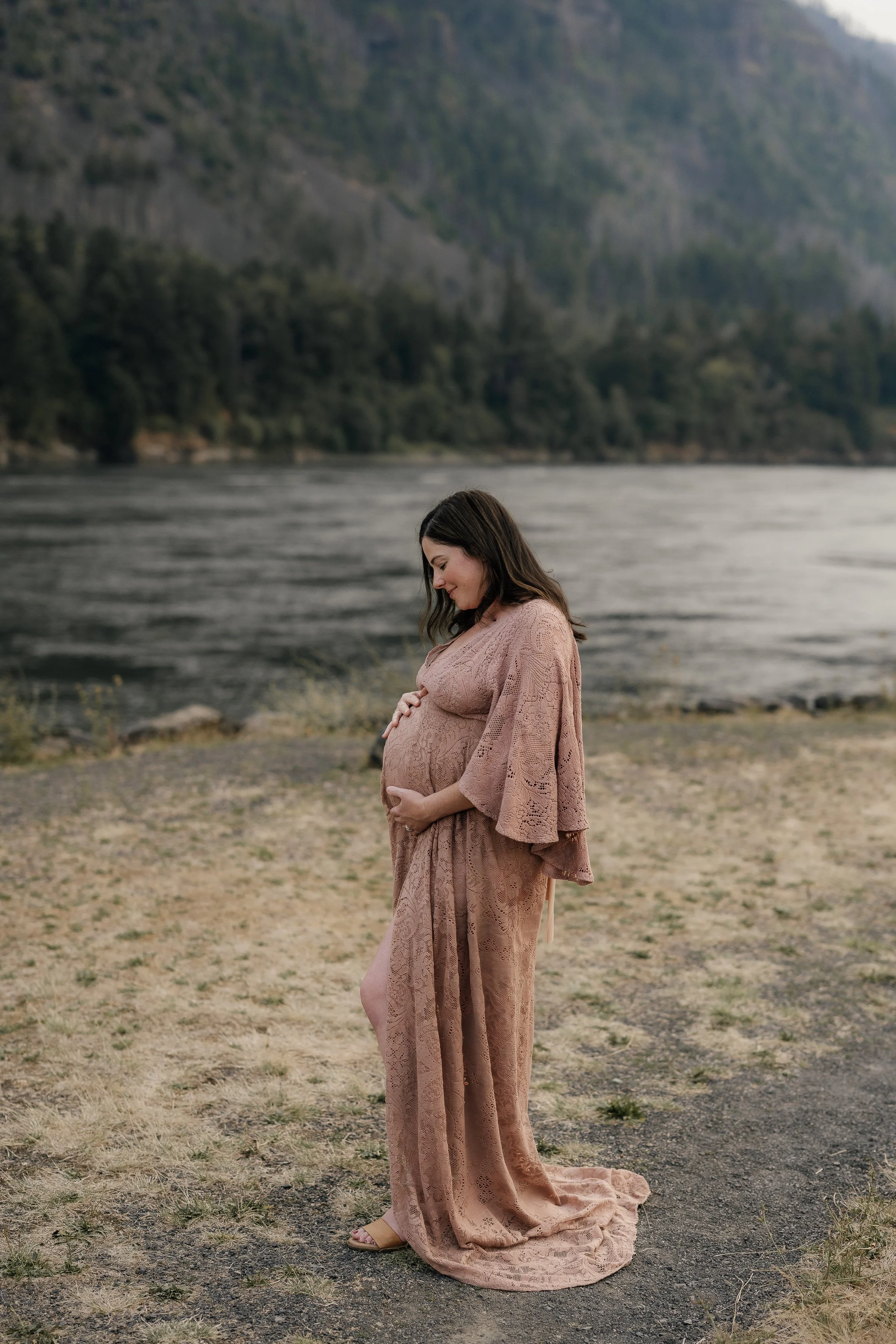 A pregnant woman in a pink lace dress stands on a grassy area near a river, holding her belly and looking down at it with a gentle smile, with a backdrop of mountains and trees.