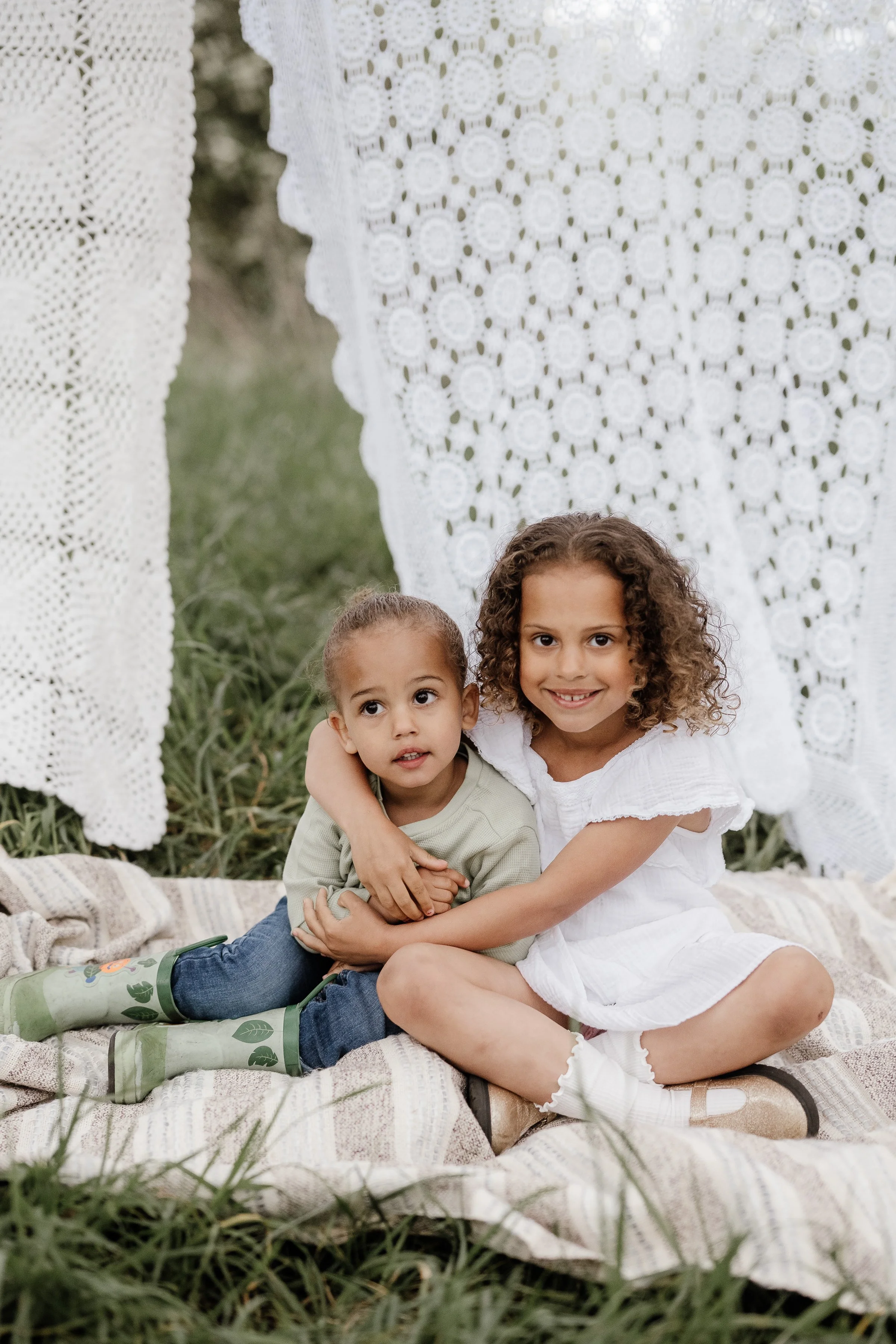 Two young girls sitting on a blanket outdoors, hugging each other with lace curtains hanging behind them.