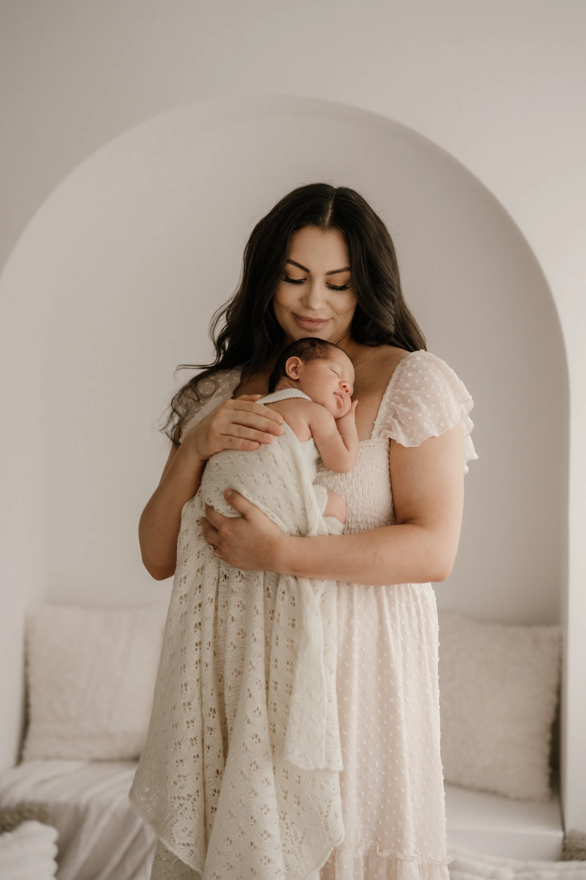 A woman in a white dress holding a sleeping newborn baby wrapped in a lace blanket, standing in a light-colored room with a minimalist aesthetic.