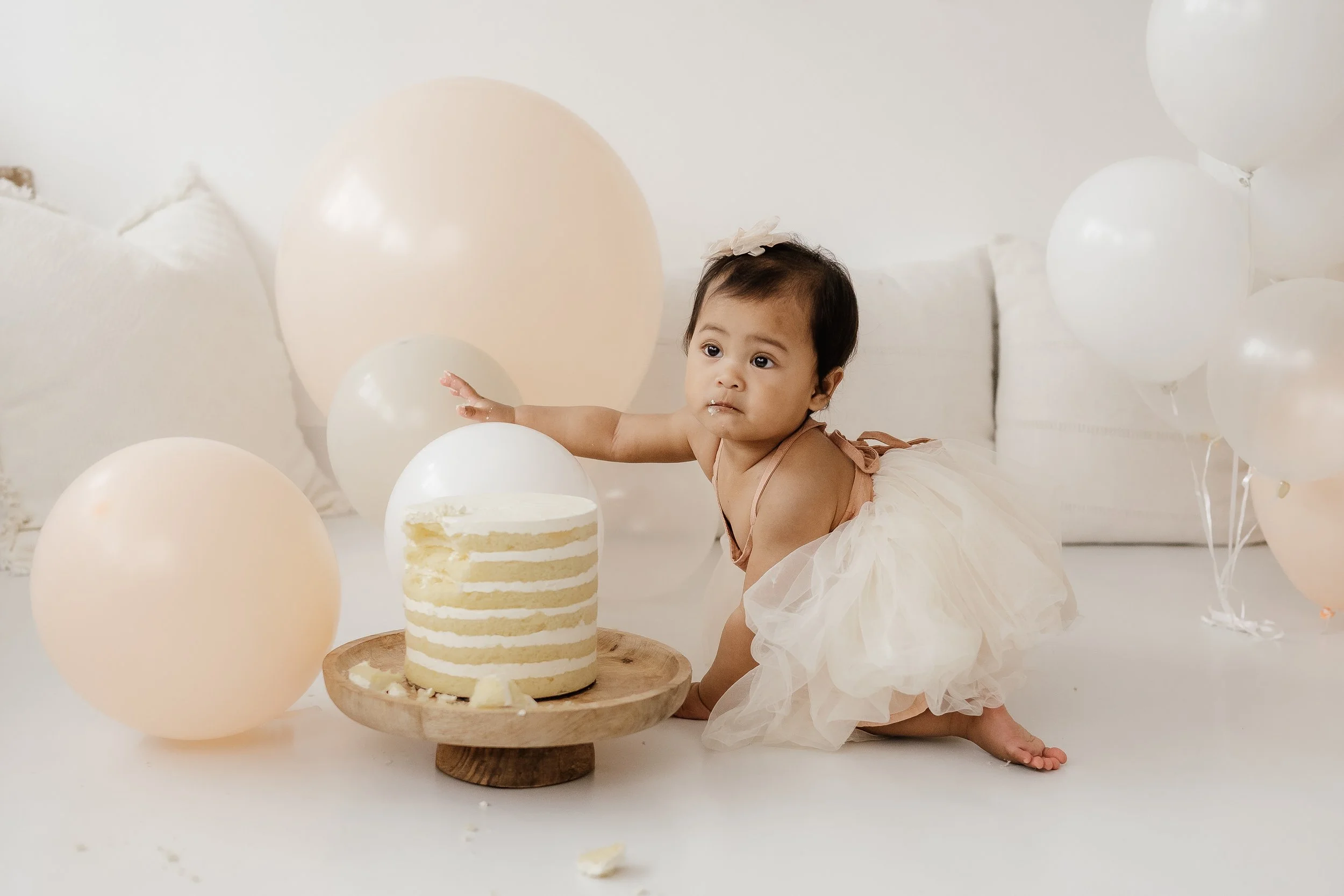Baby girl in a cream-colored tutu reaching for a cake with white frosting and multiple layers, surrounded by cream and peach balloons in a white room.