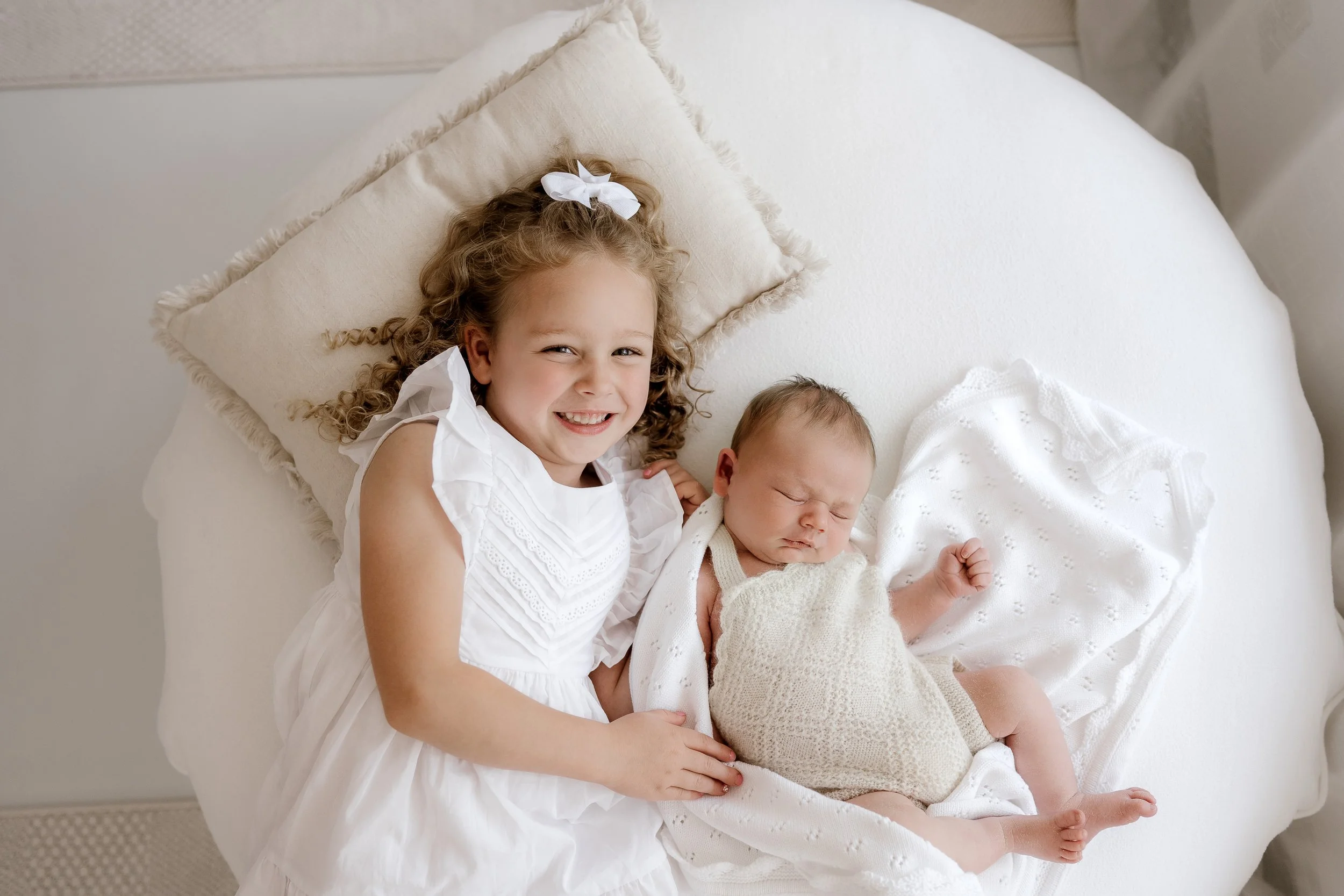 A young girl with curly hair and a white dress lying on a bed next to a newborn baby who is sleeping, swaddled in a blanket.