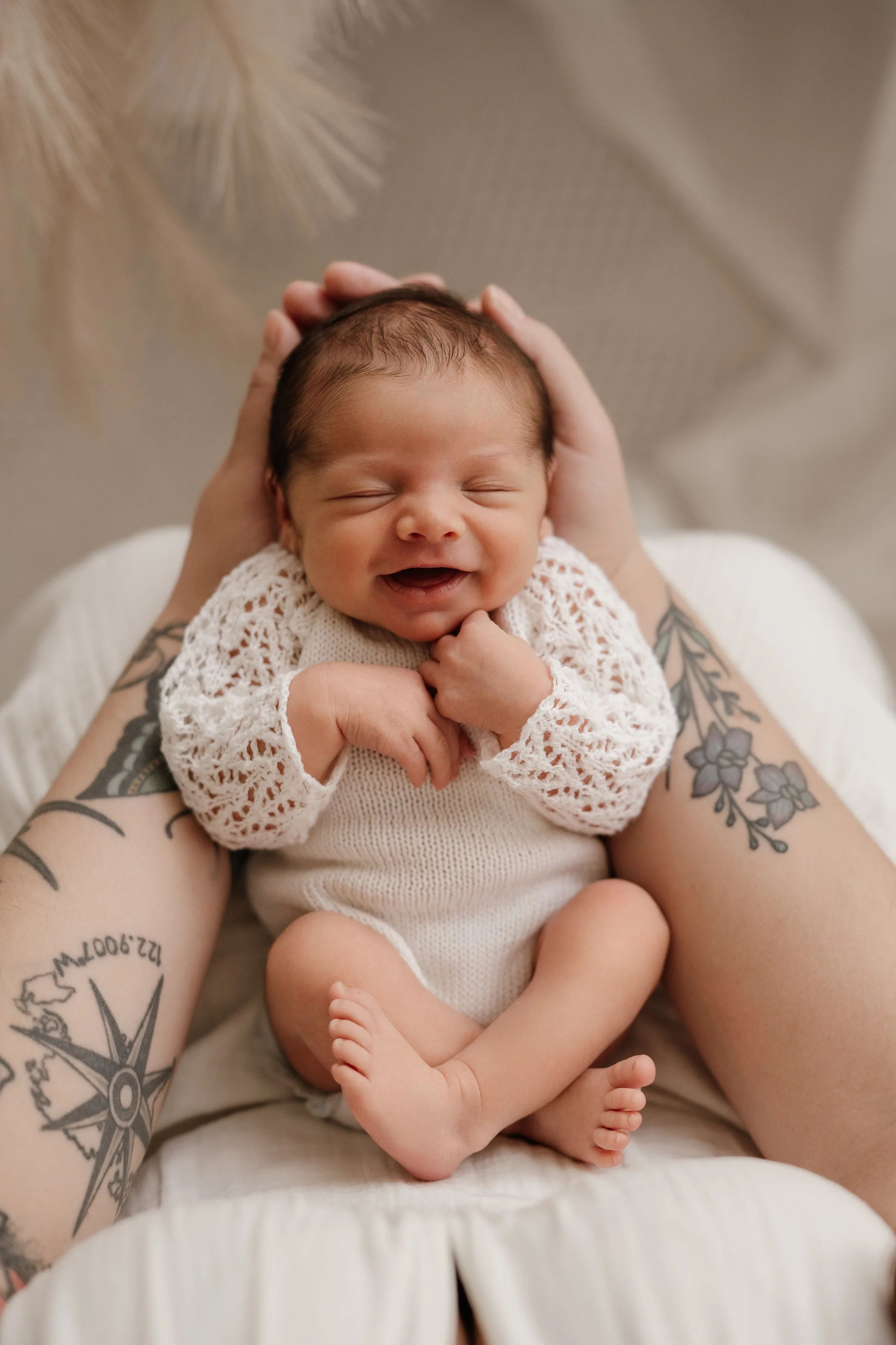 A smiling newborn baby with closed eyes, wearing a white knitted sweater, resting on a person's lap with tattoos, who is gently holding the baby's head.