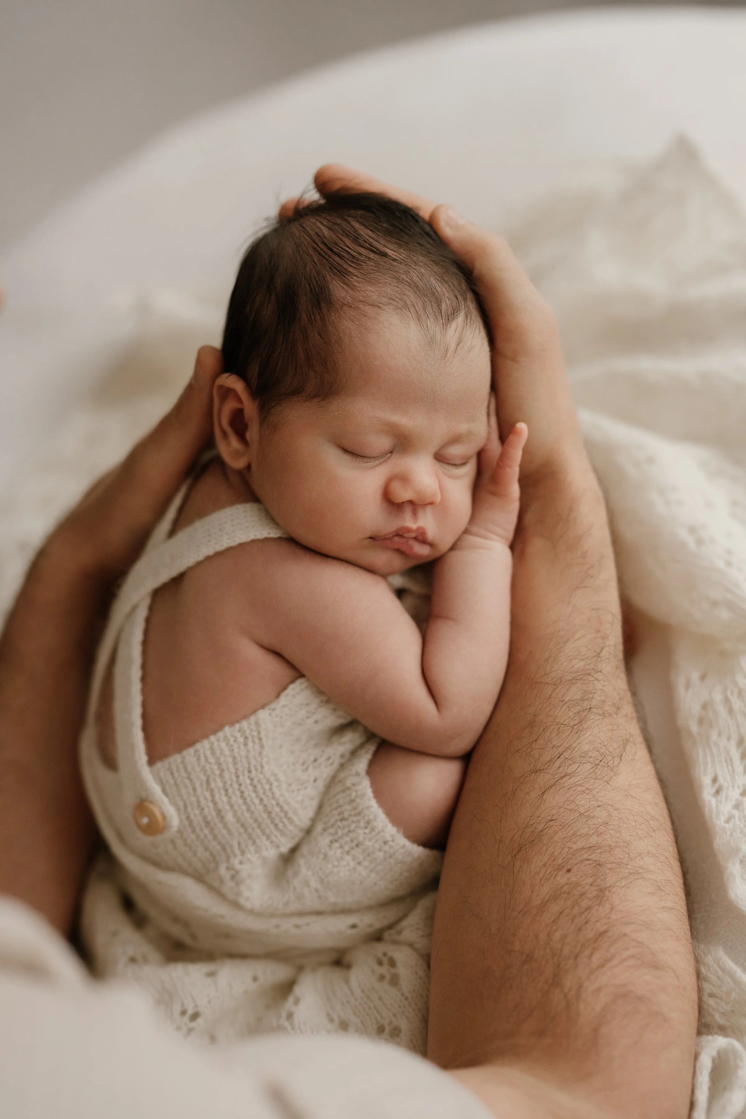 Close-up of a tender moment with a sleeping baby resting on an adult's arms, showing the baby's peaceful face and arm curled with fingers touching their cheek, all wrapped in a cozy knitted outfit.