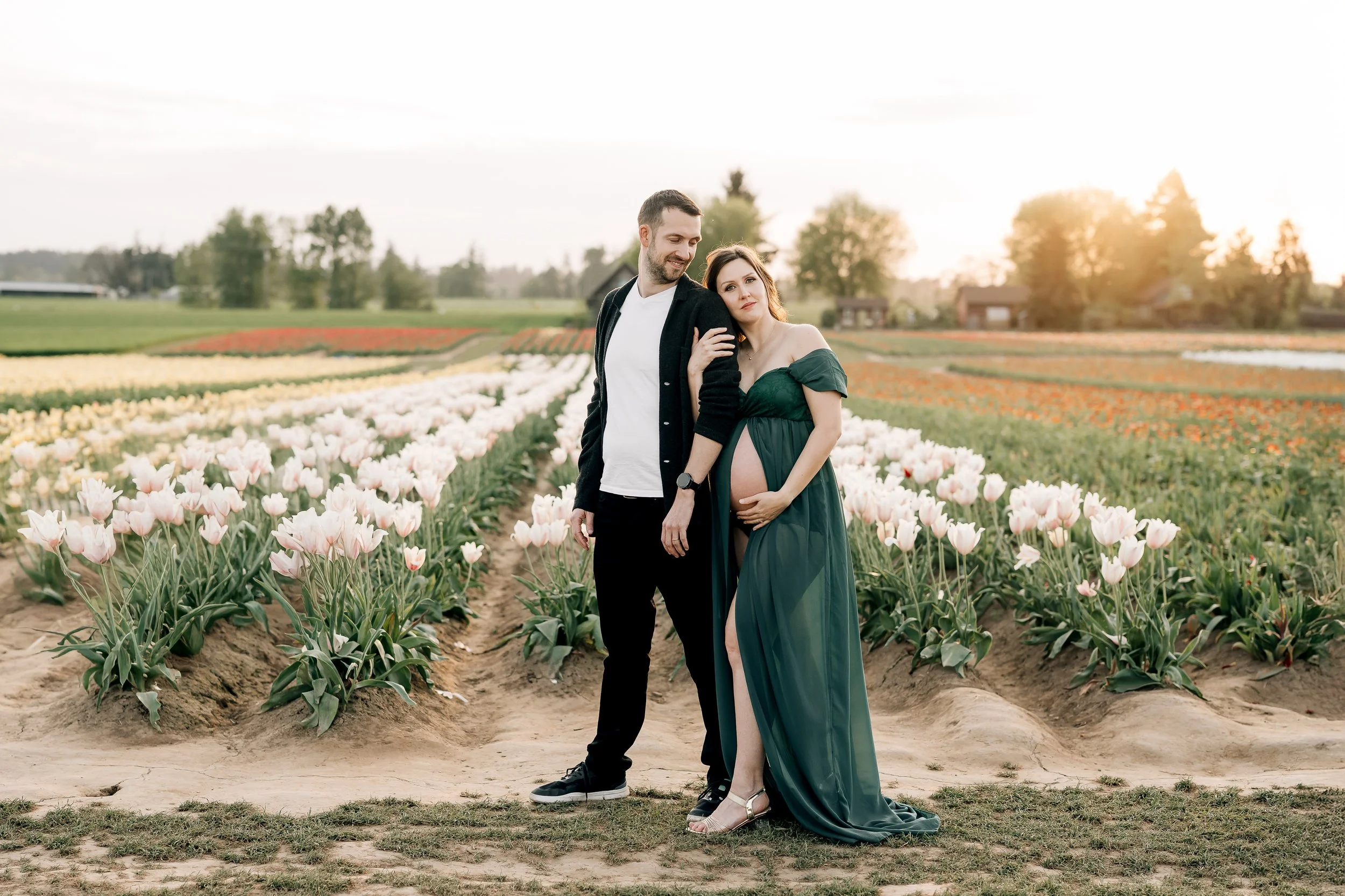A pregnant woman in a green dress with a slit, standing next to a man in black pants and a black jacket, in a tulip field during sunset.