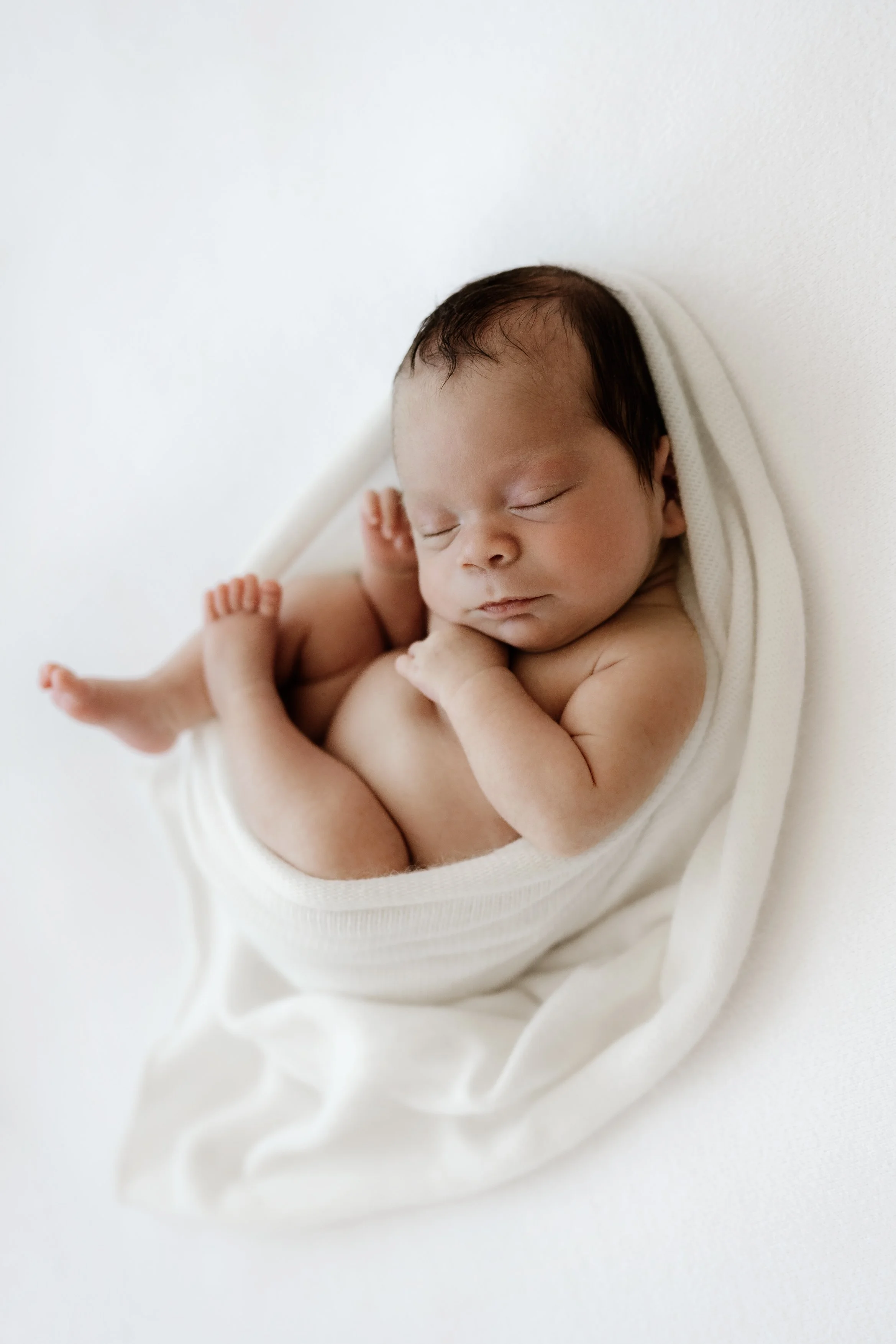 A sleeping newborn baby swaddled in white cloth, lying on a white surface.