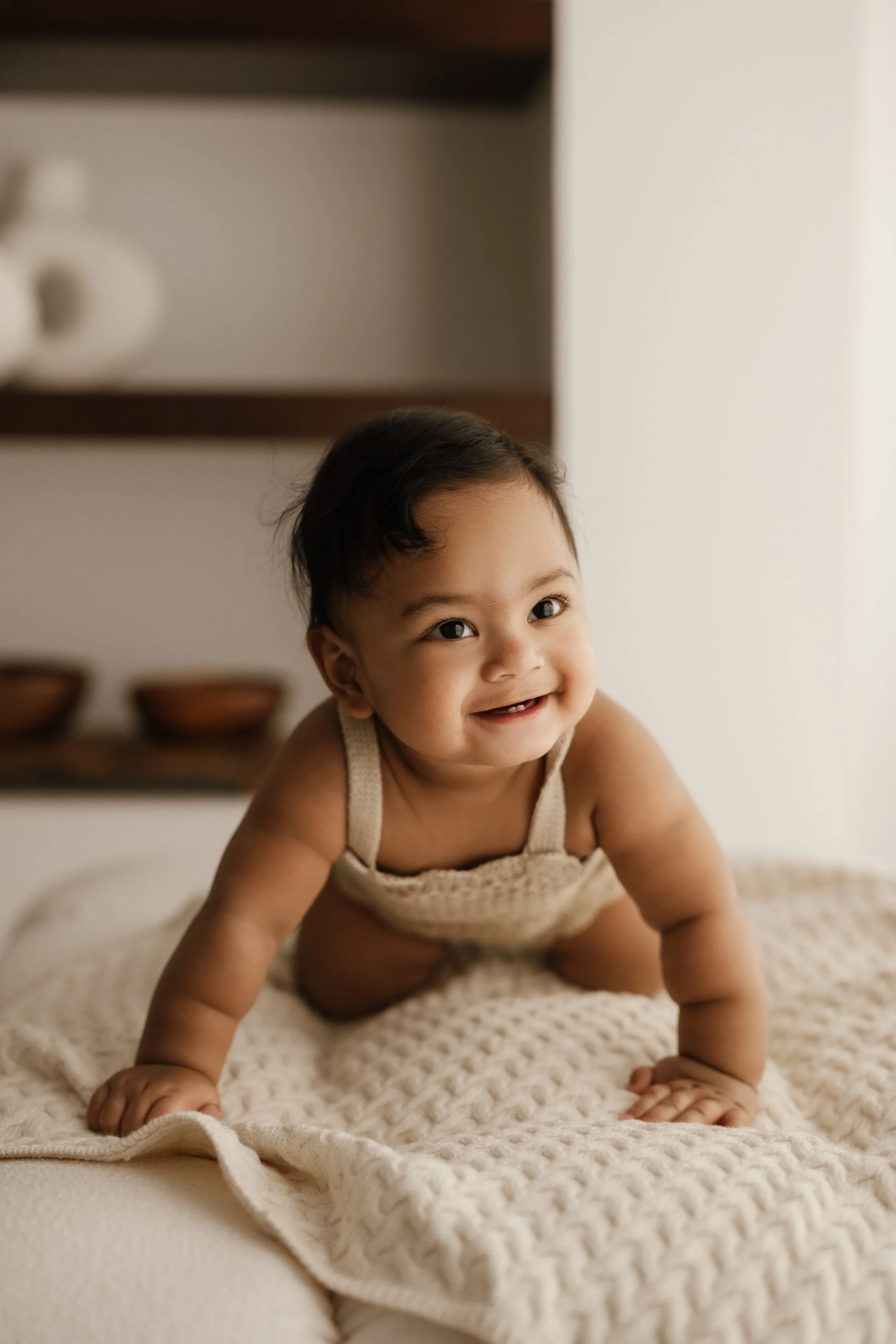 Smiling baby crawling on a cream-colored blanket in a cozy, softly lit room.