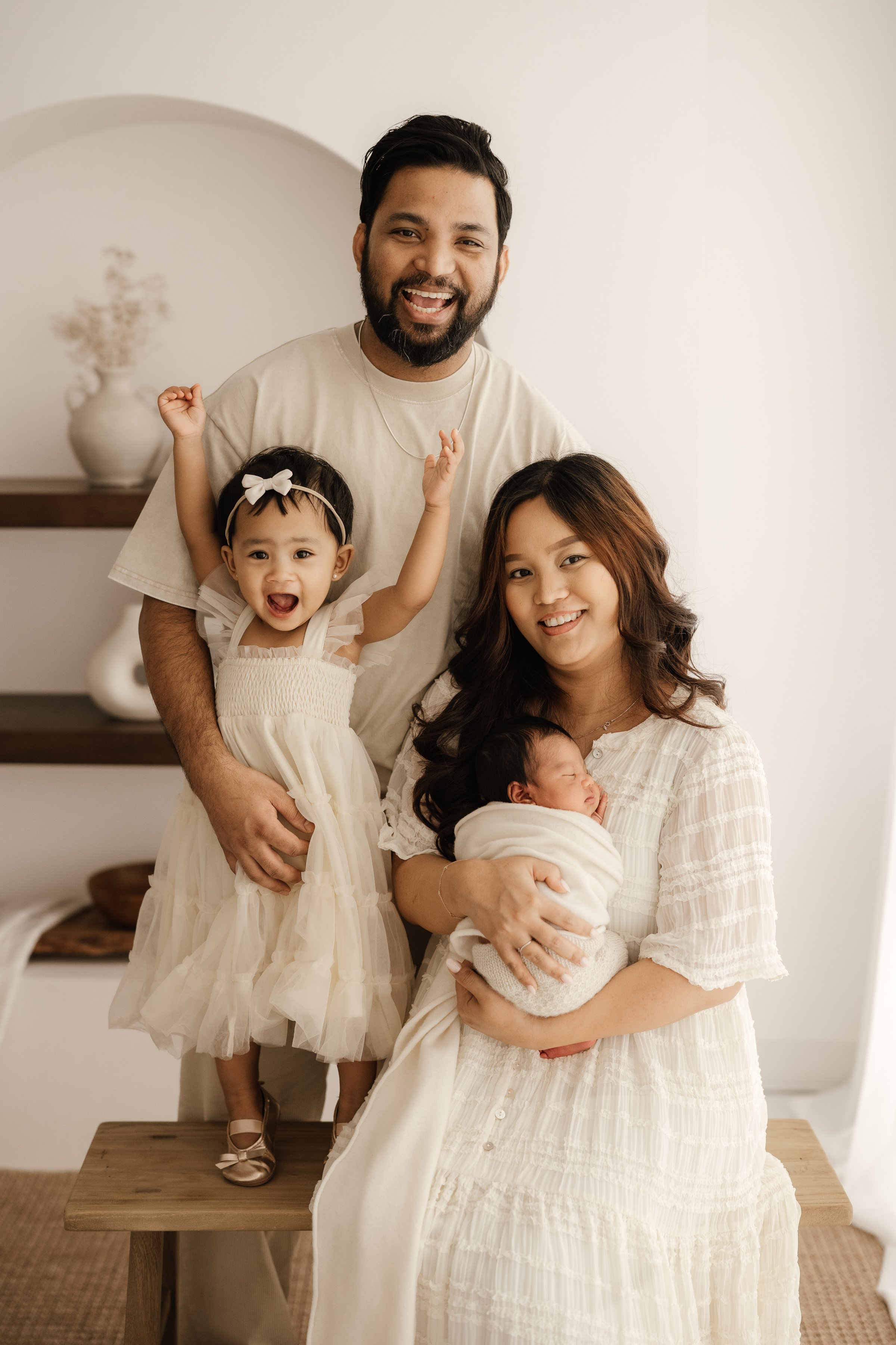 Family gathering with two women, a man, a toddler girl, and a newborn baby in a cozy home setting.