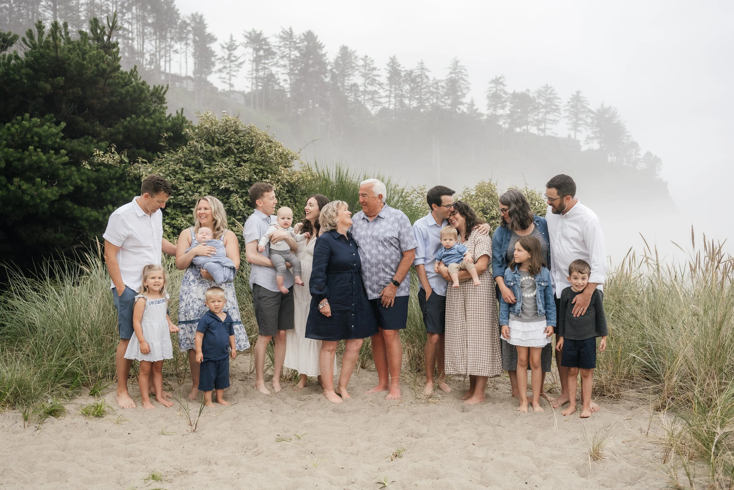 A large multigenerational family standing on a sandy beach with grass and trees, smiling and enjoying each other's company.