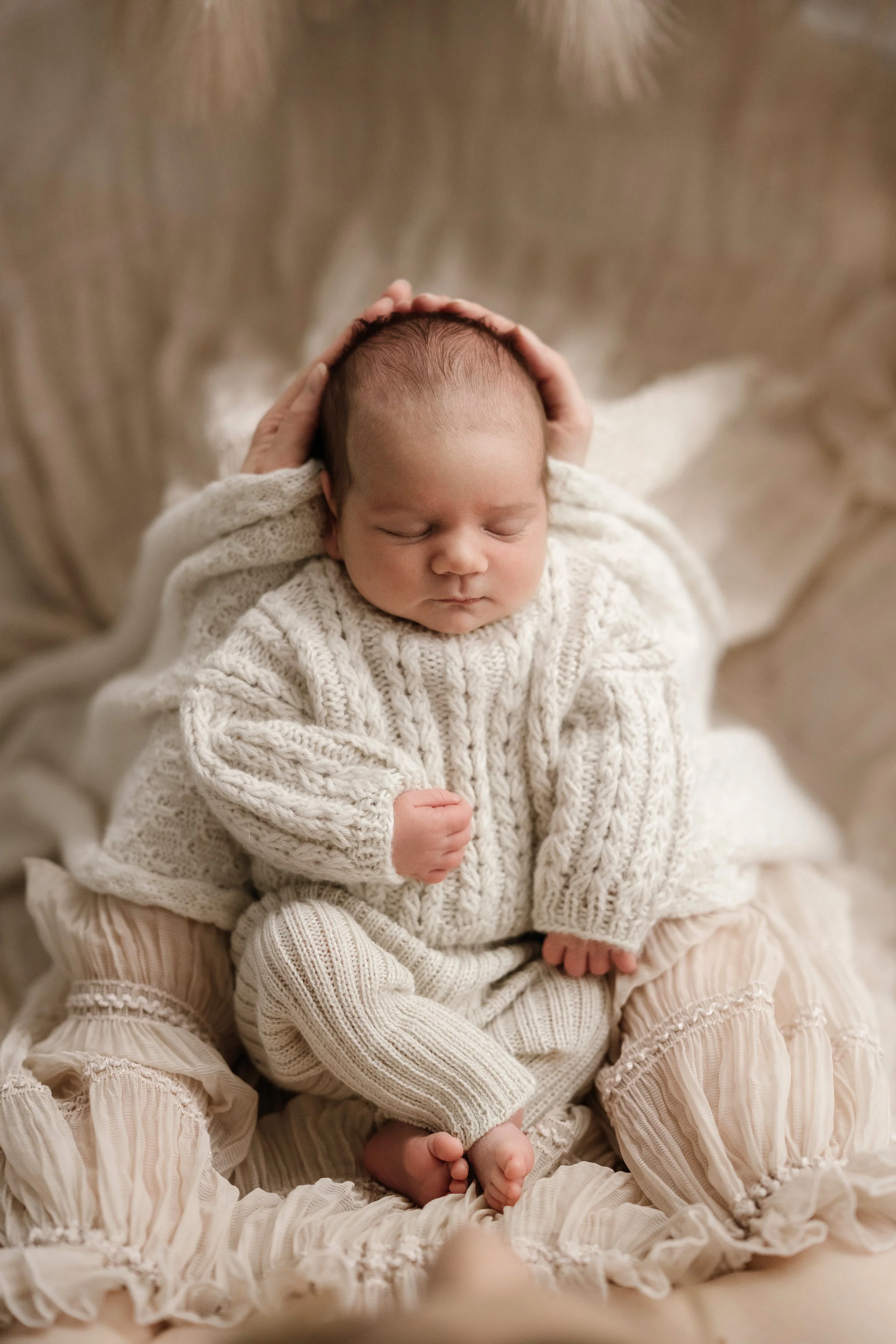 A sleeping newborn baby dressed in cream-colored knit clothing, cradled gently in an adult's lap, with a soft, cozy background.