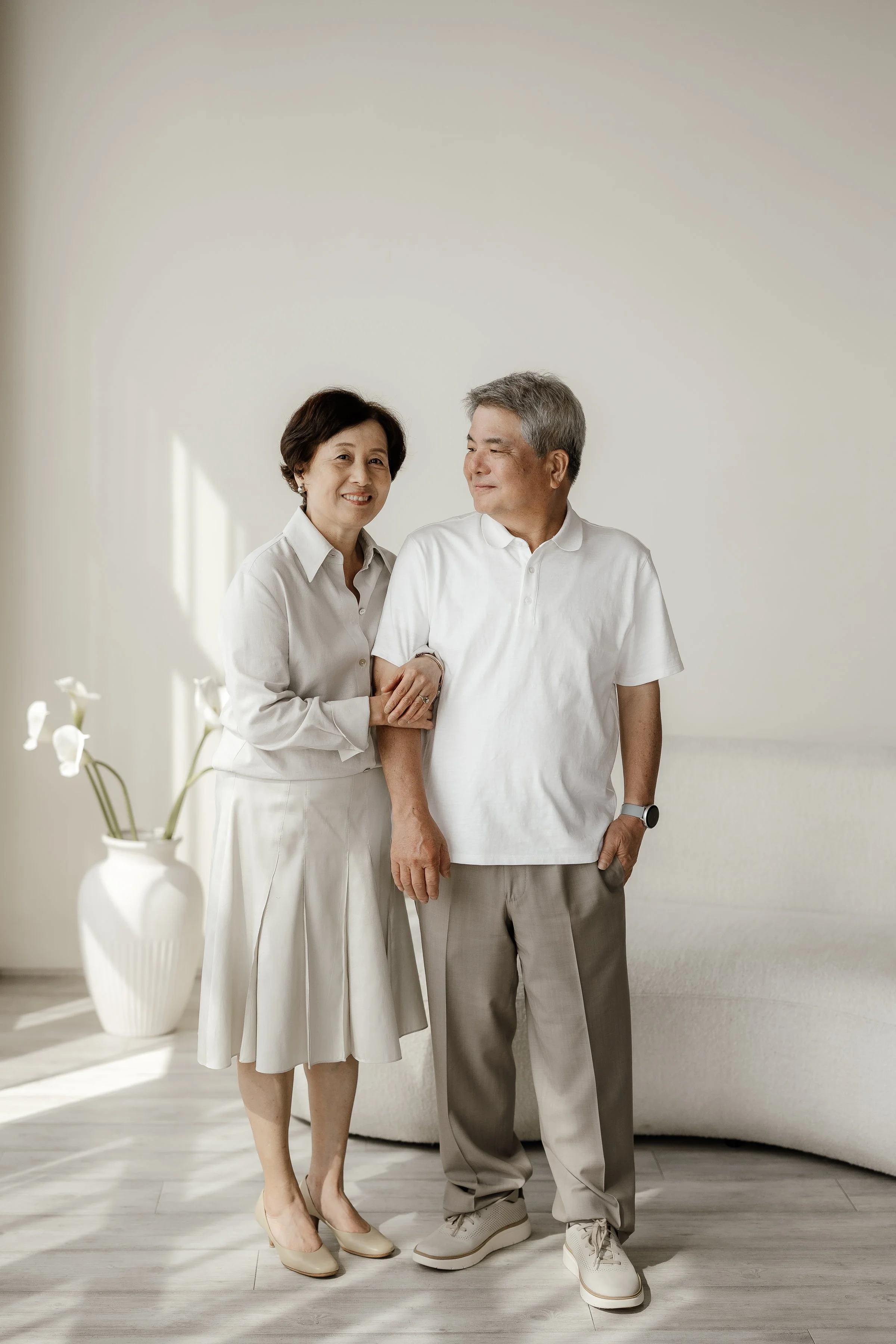 An elderly couple smiling and happily standing together indoors, with a minimalistic background and a white flower vase with lilies nearby.