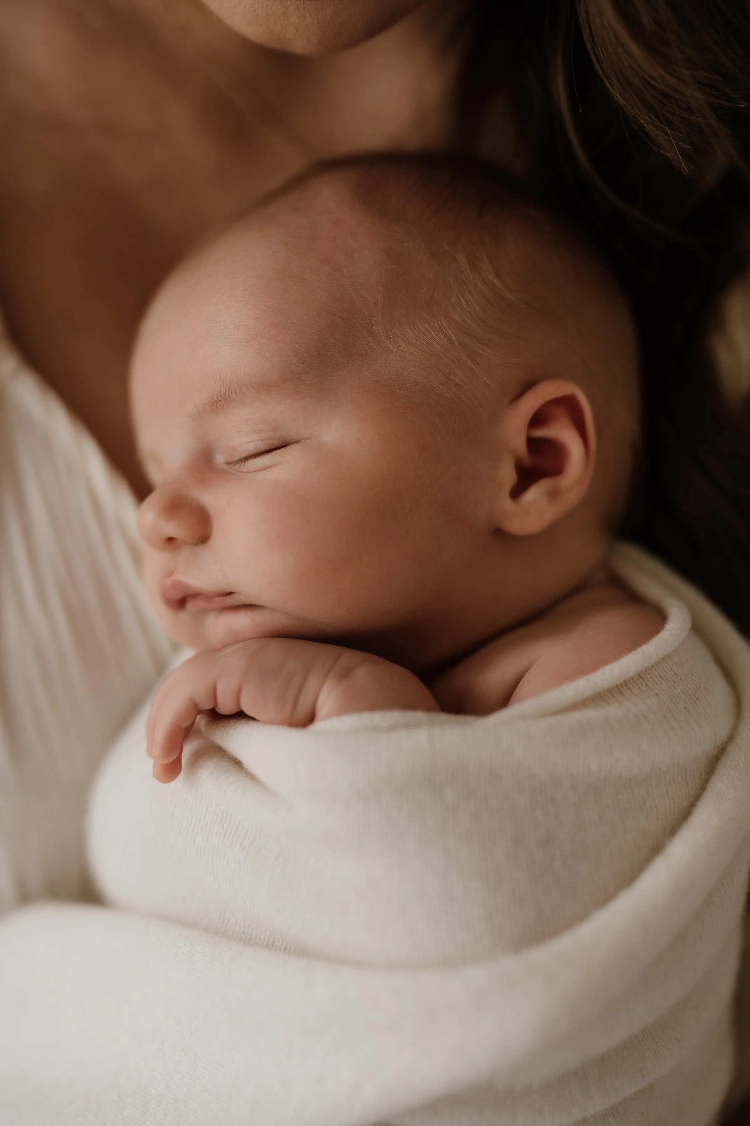 Close-up of a sleeping baby with a peaceful expression, resting on someone's shoulder, wrapped in a soft cream-colored blanket.