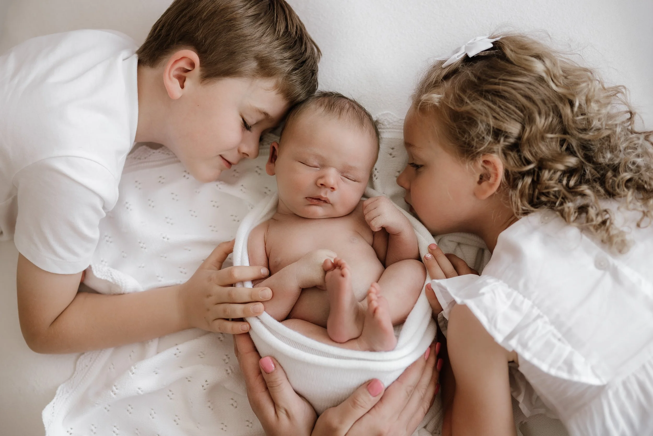 Two children, a boy and a girl, gently holding and looking at a sleeping baby wrapped in a white blanket.