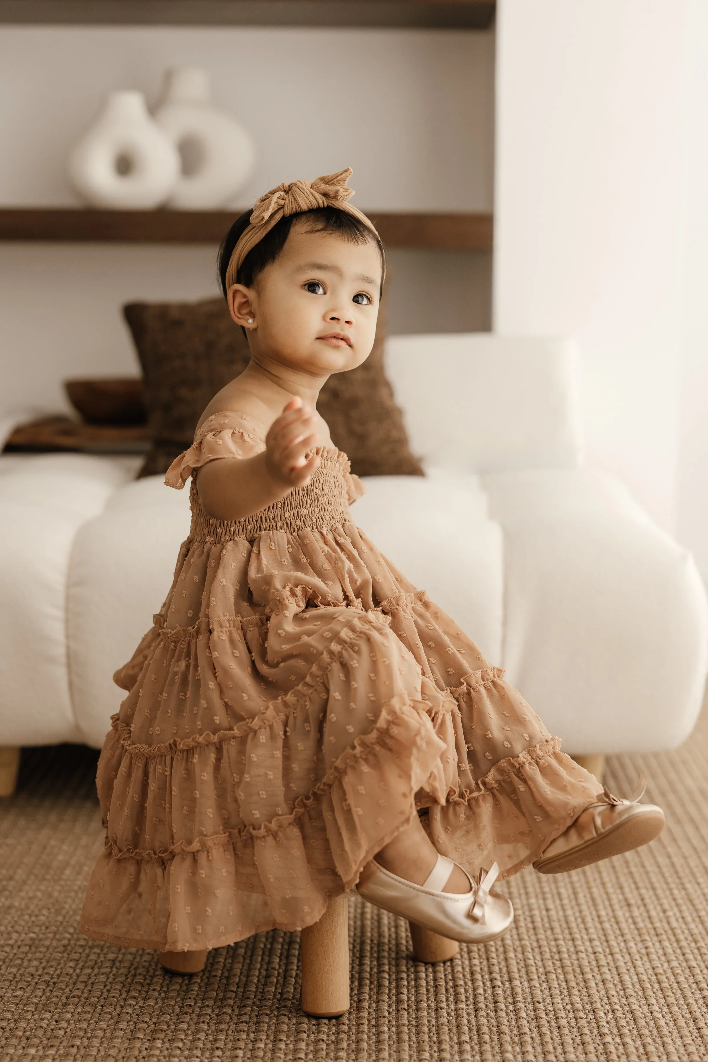 Young girl sitting on a small wooden stool in a living room, wearing a beige dress with ruffles and polka dots, a matching headband, and cream-colored shoes with bows.