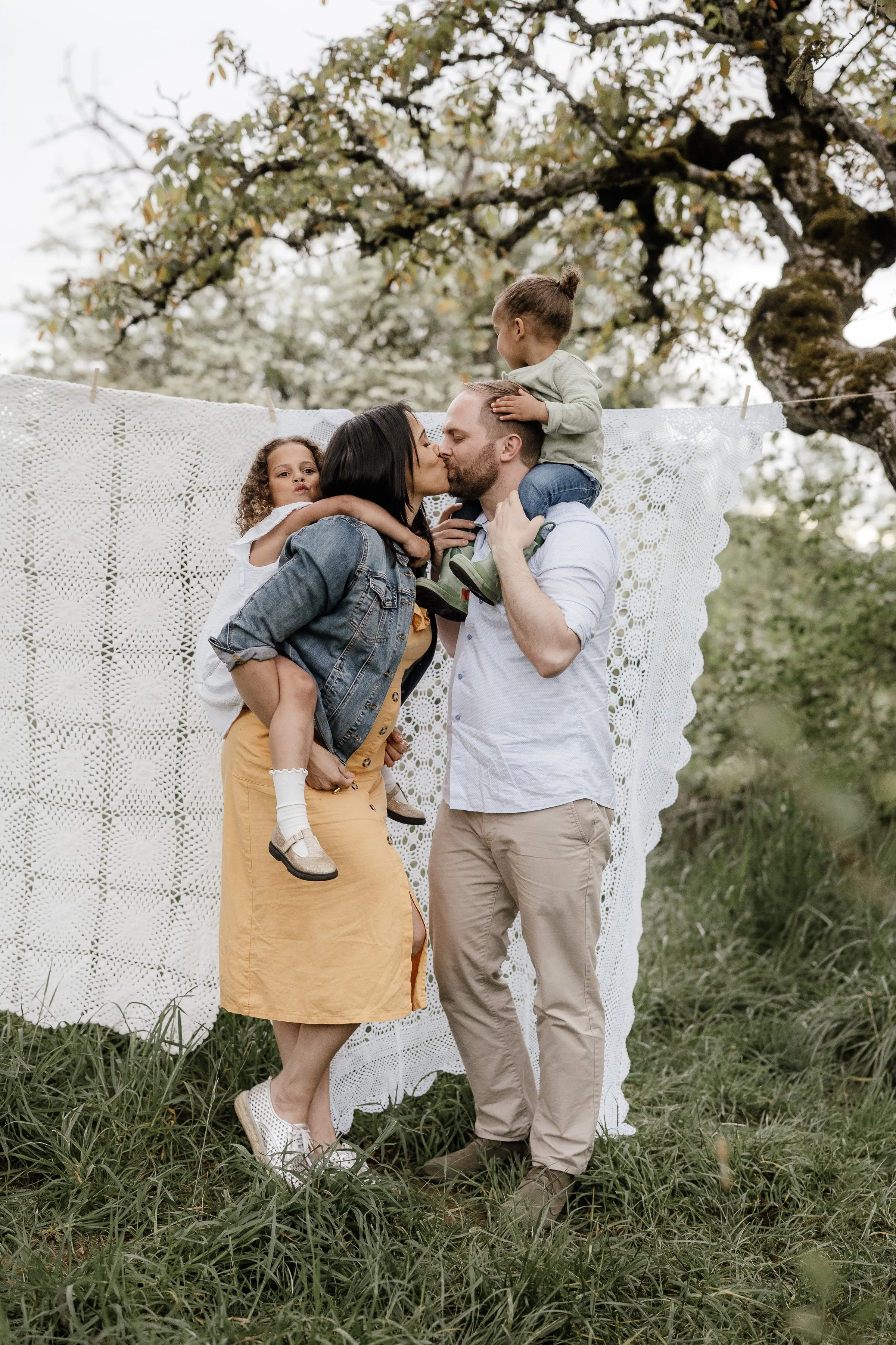 A family of four sharing a kiss outdoors, with a young girl on her mother’s back, a young boy sitting on his father’s shoulders, standing in front of a white lace blanket hanging from a tree.