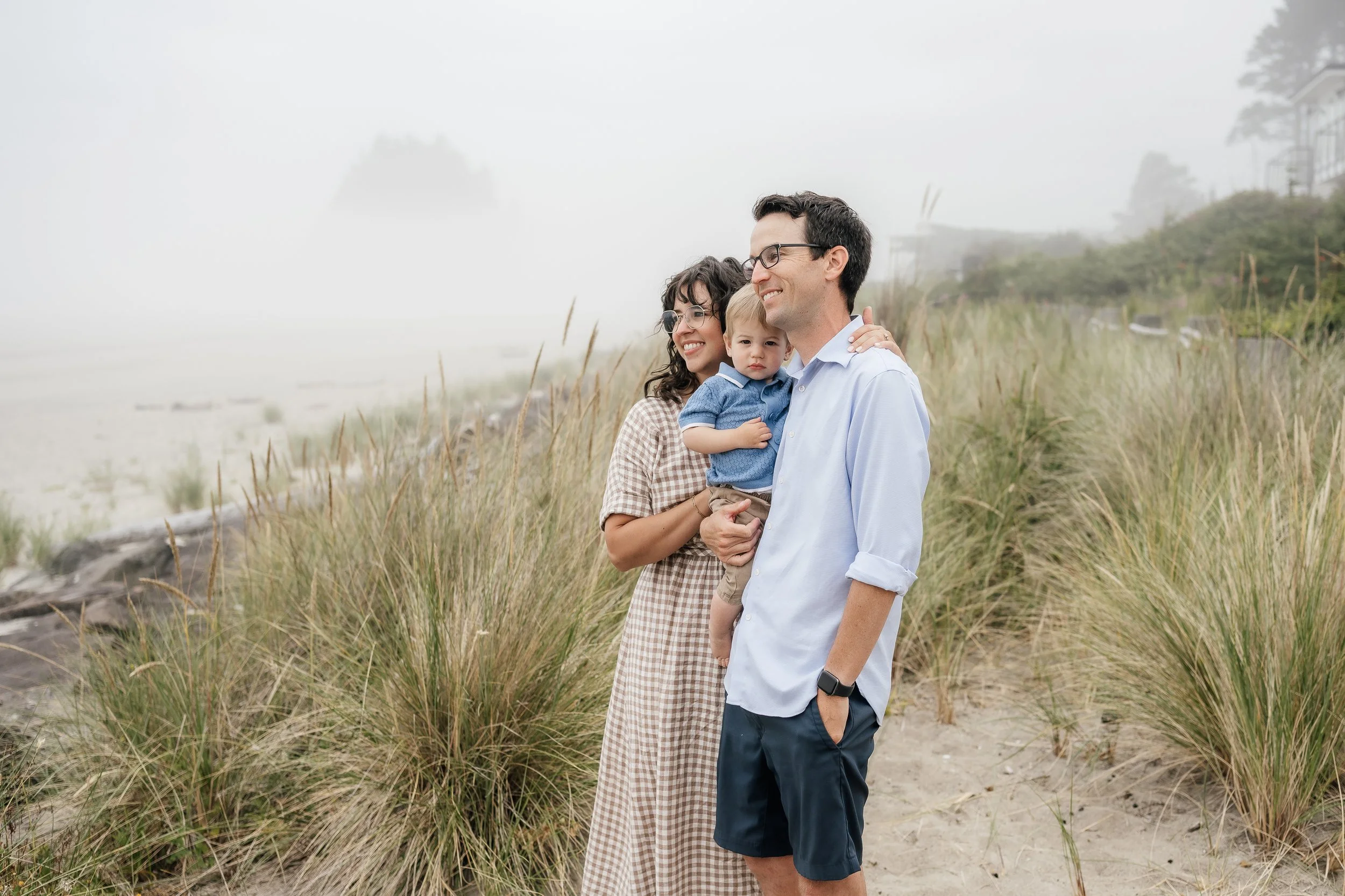 A family of three standing on a foggy beach with tall grass, smiling and hugging, with the mother and father holding their young son.