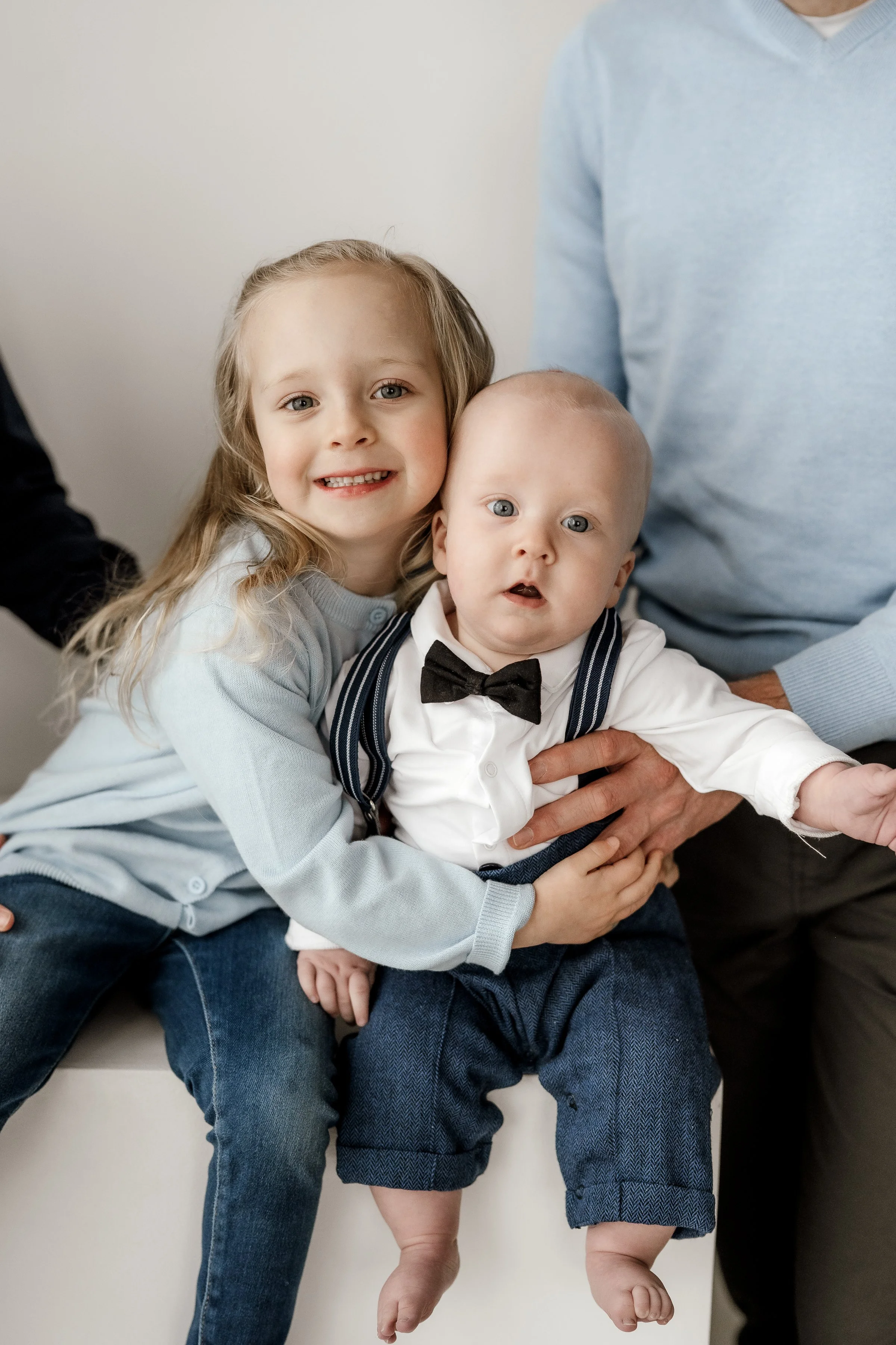 A young girl and a baby boy dressed in formal clothes, sitting together with an adult partially visible, in a cozy indoor setting.