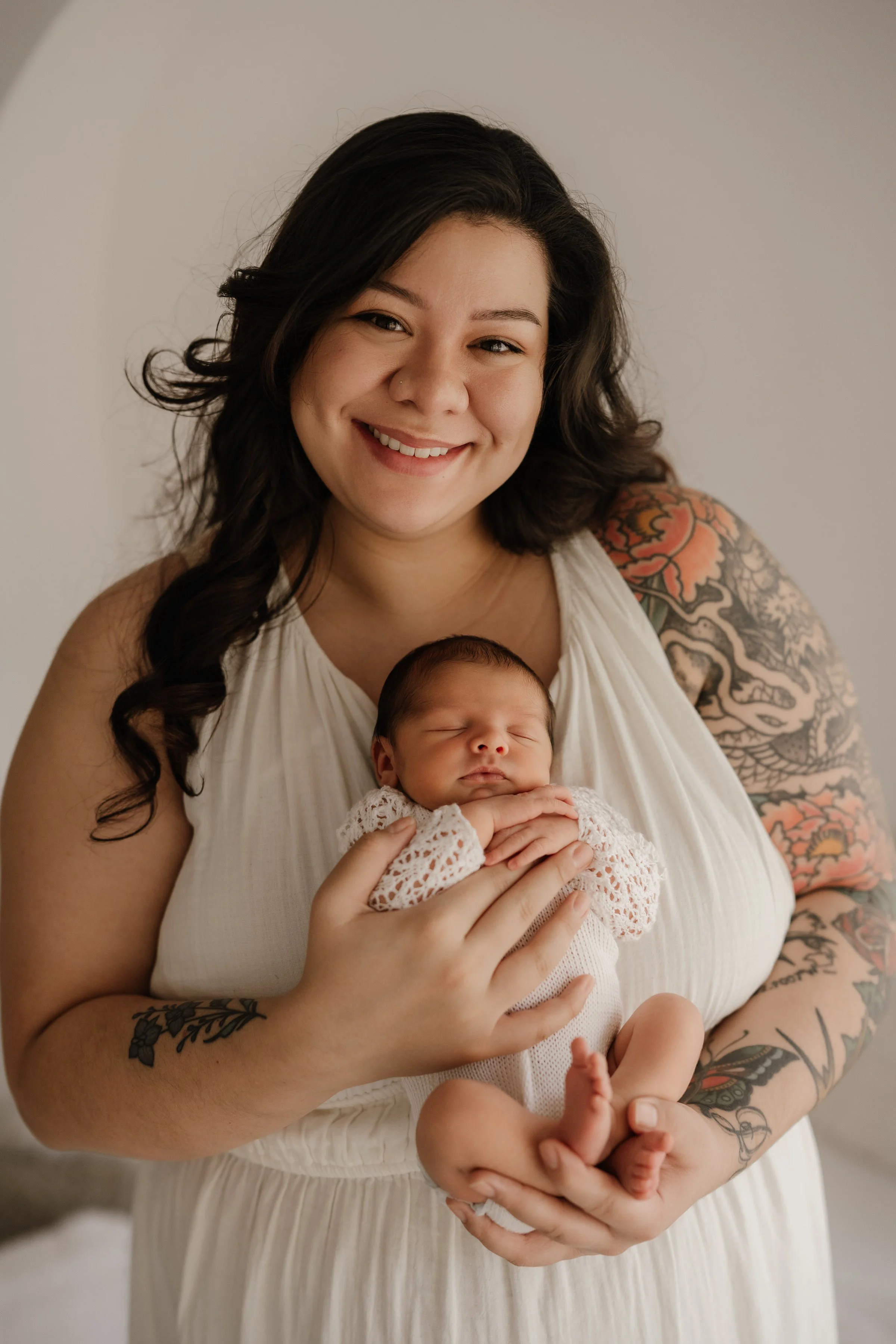 A smiling woman with dark curly hair holding a sleeping baby dressed in a white knitted outfit.