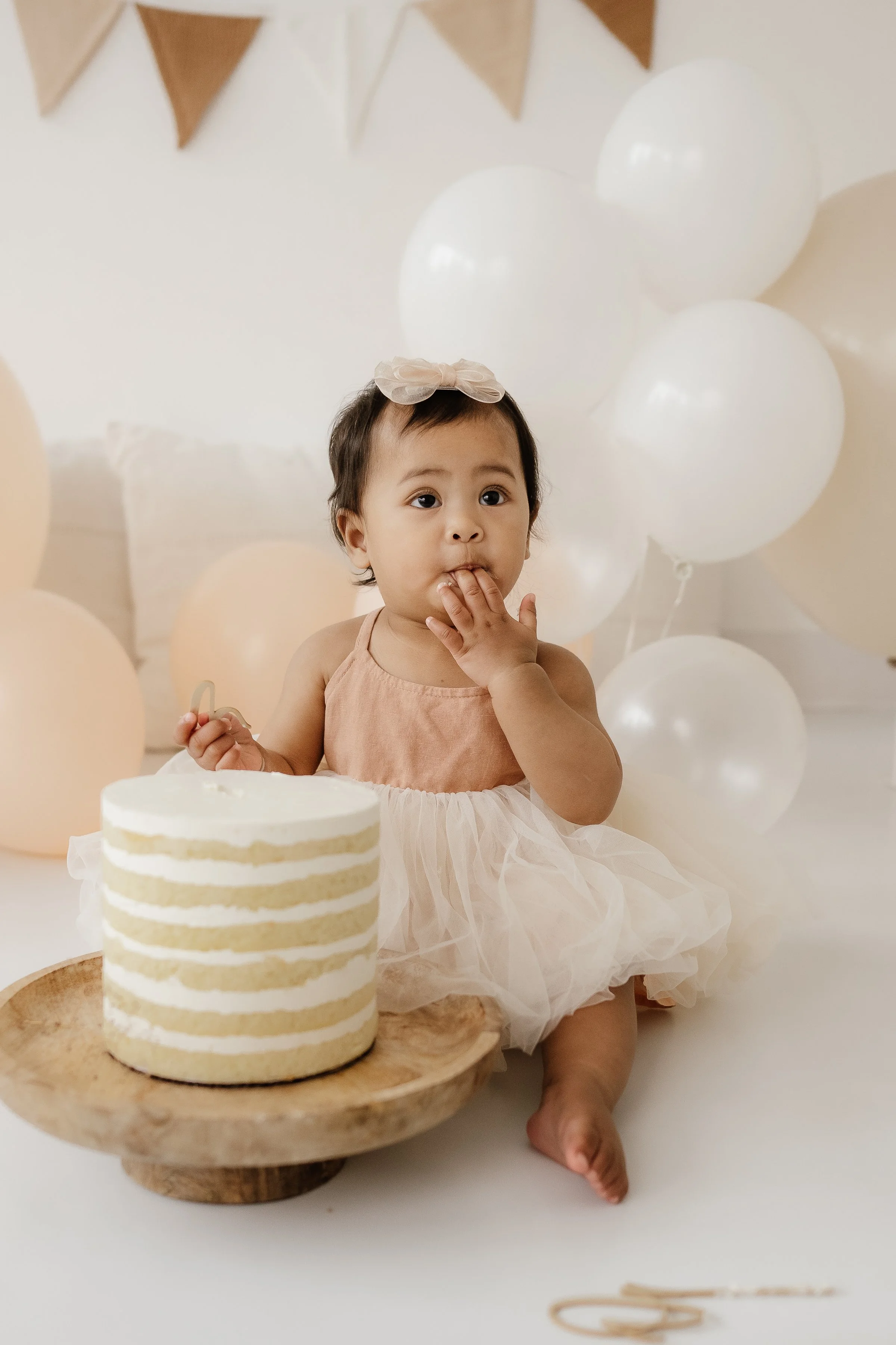 A young girl in a peach dress and tulle skirt sitting on the floor next to a birthday cake with white frosting and sponge layers, surrounded by white and beige balloons, in a decorated room.
