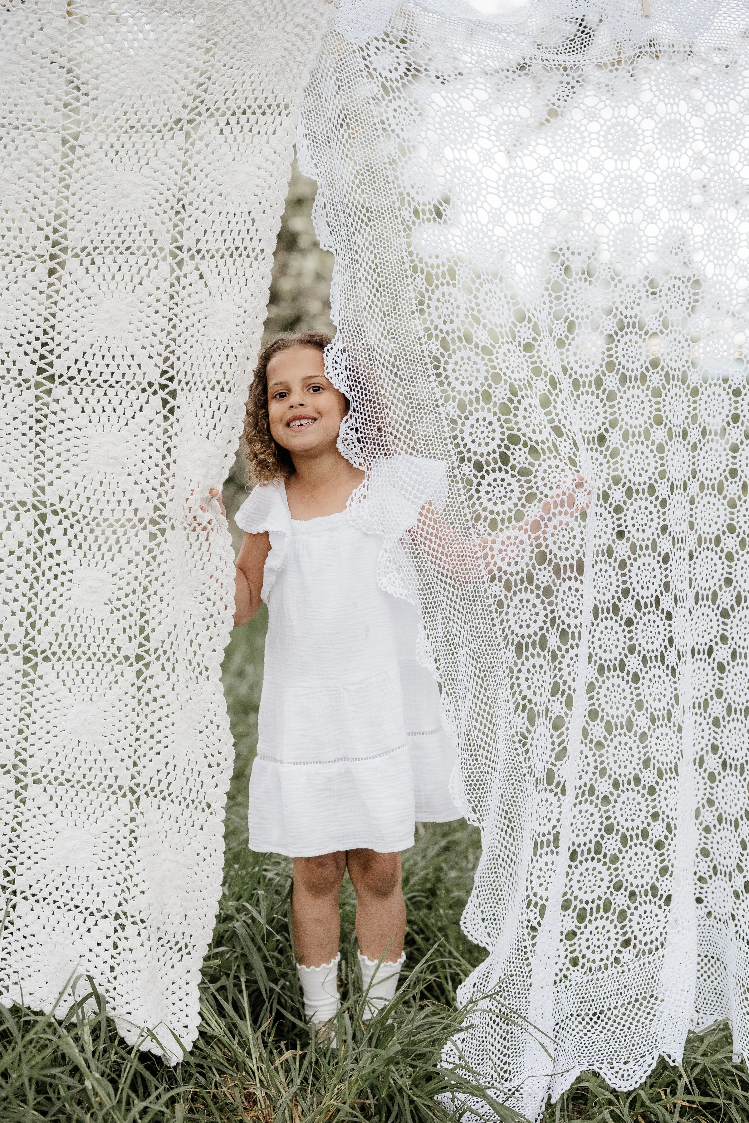 Young girl with curly hair wearing a white dress and socks, smiling, standing behind and peeking through lace curtains outdoors.