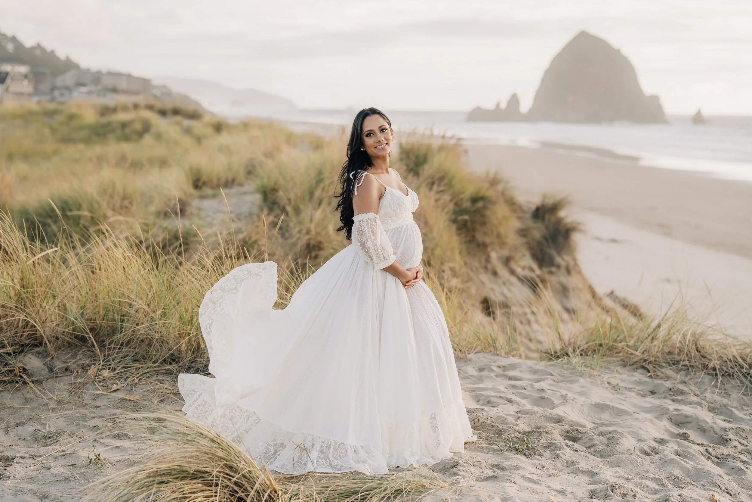 Pregnant woman in a white flowing dress standing on a sandy beach with grassy dunes and a large rock formation in the background.