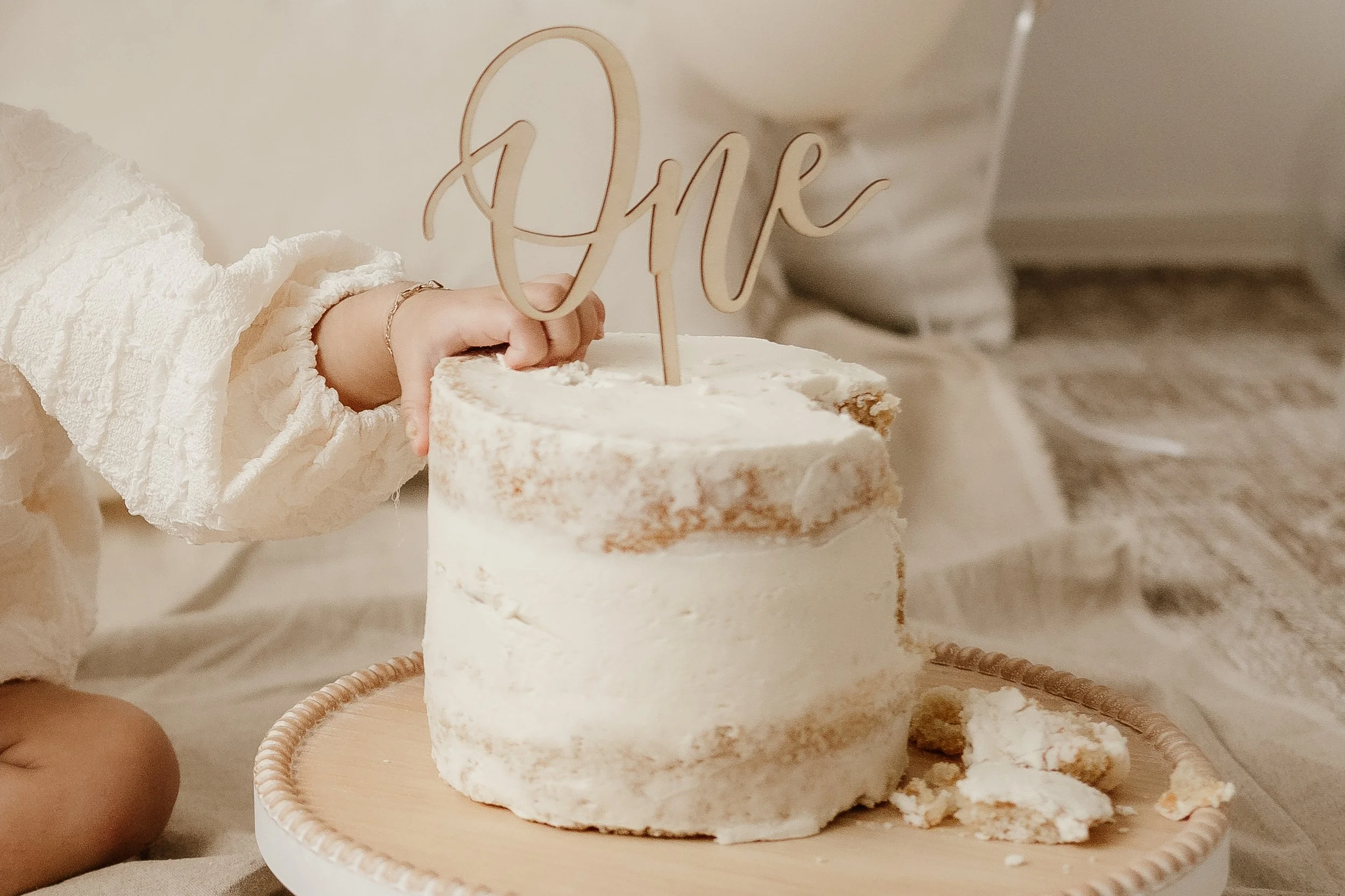 Child's hand holding a piece of a white frosted birthday cake with a gold 'One' cake topper, on a round wooden cake stand, in a cozy indoor setting.