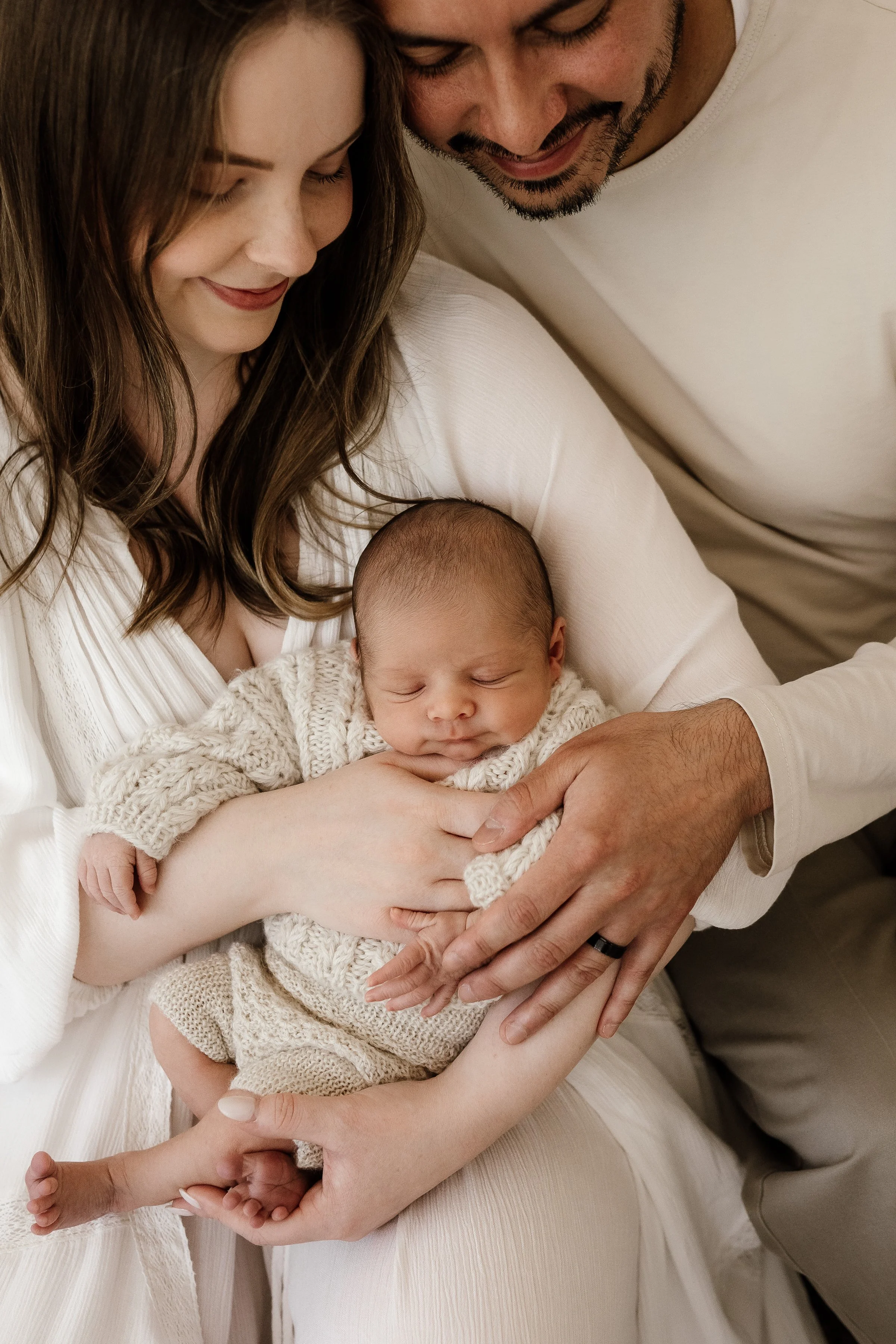 A young family holding their sleeping infant, with the mother and father smiling and looking at the baby, dressed in neutral-colored clothing.