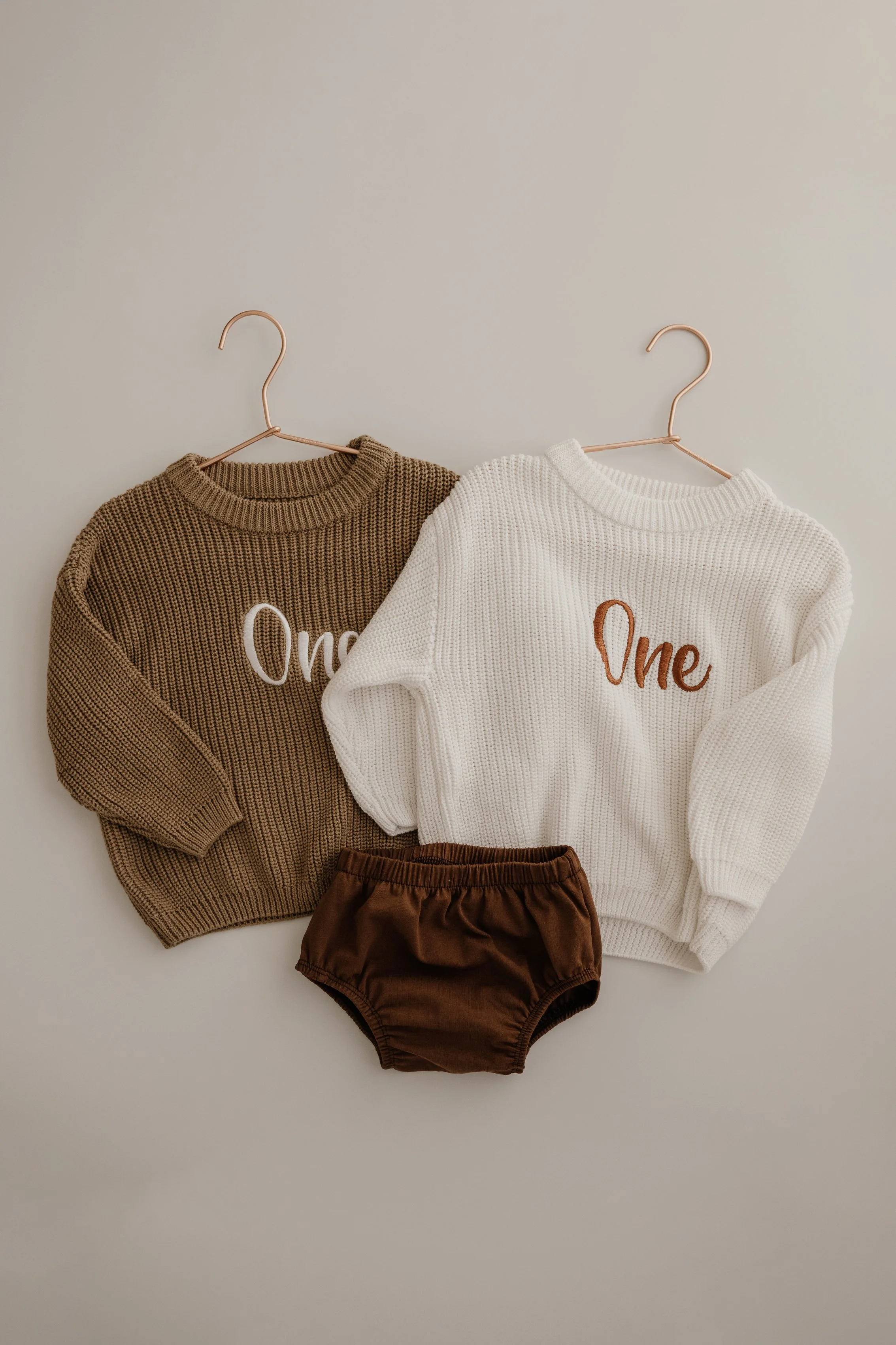 Two children's sweaters, one brown and one white, hanging on pink hangers, with a pair of brown baby shorts placed in front of them on a plain white background.