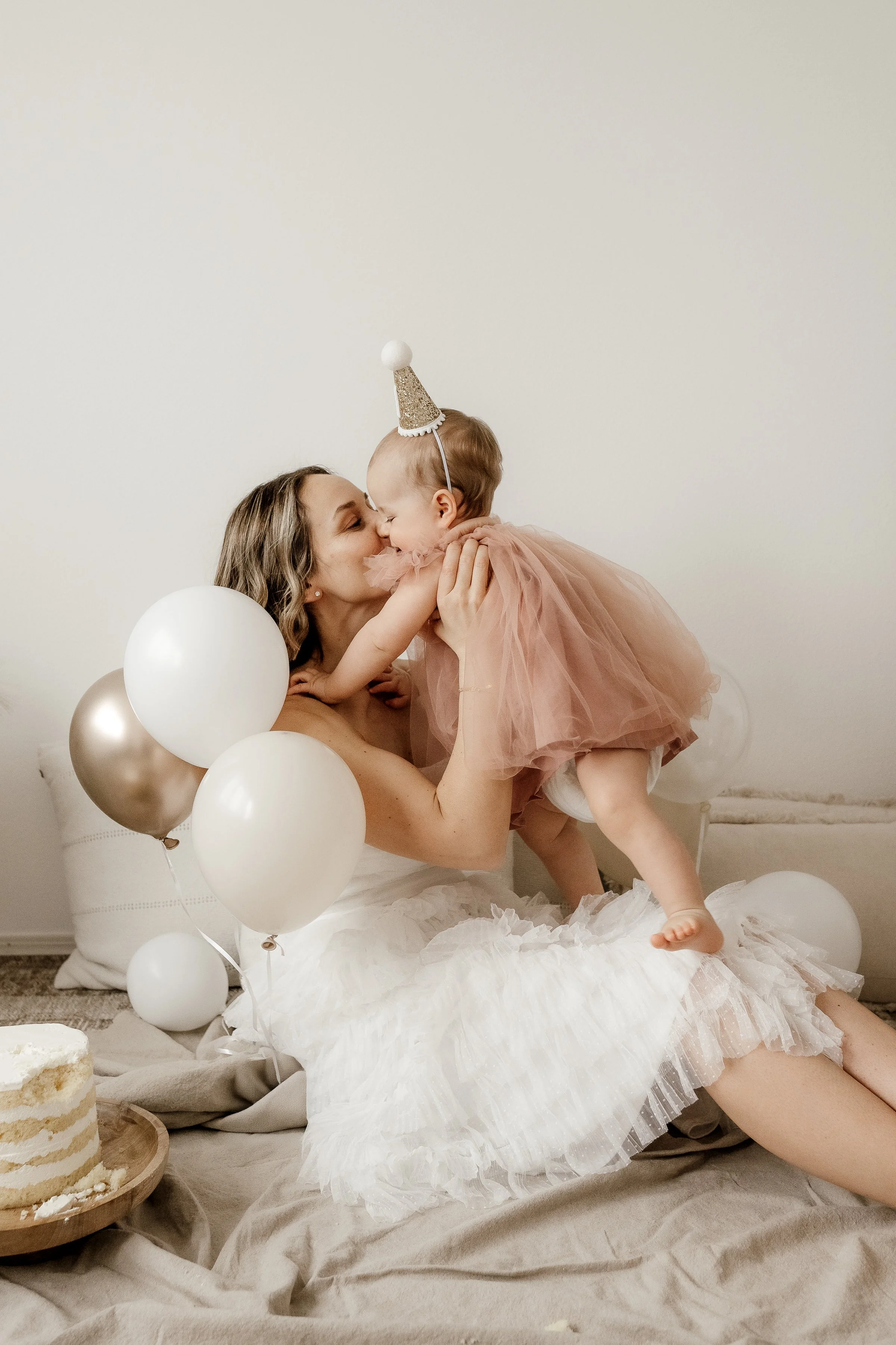 Mom and young daughter celebrating a birthday with balloons and cake, girl wearing a party hat and pink tulle dress, mom kissing daughter, cozy indoor setting.