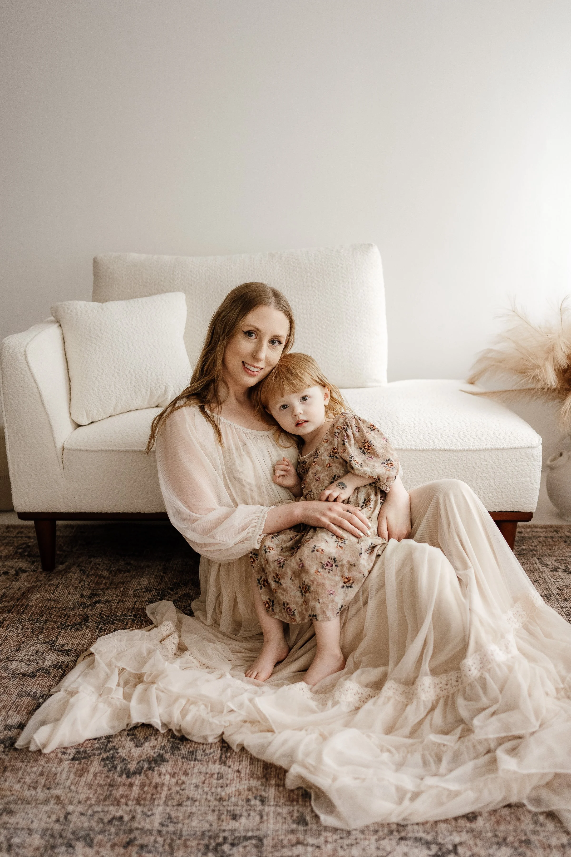 A woman and a young girl sitting on the floor in front of a white sofa, with the woman holding the girl, both wearing light-colored dresses, in a cozy living room.
