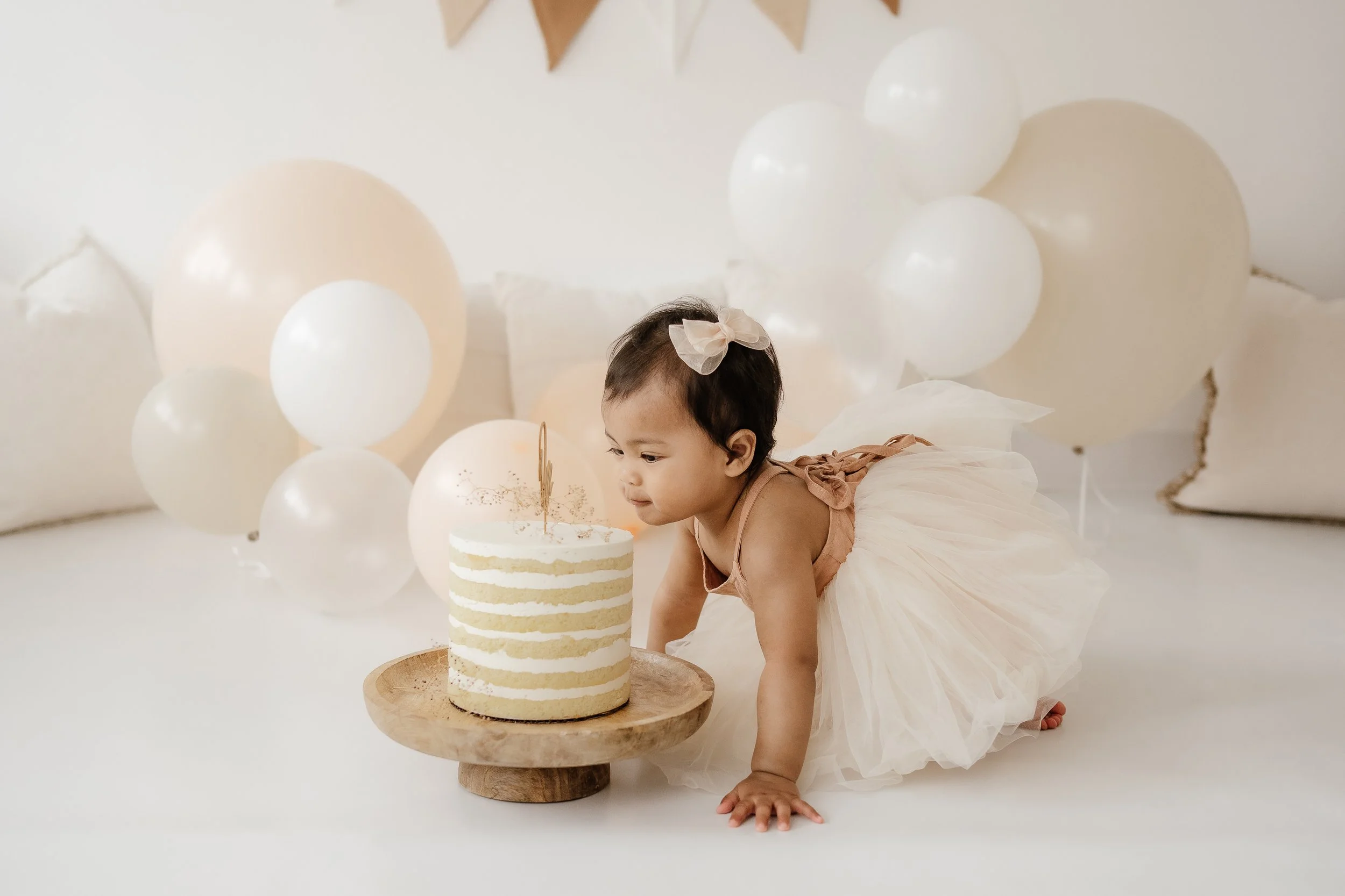 Young girl in a peach dress and tutu, with a bow in her hair, crawling on a white surface next to a layered birthday cake on a wooden stand, surrounded by white and beige balloons in a room with neutral decor.