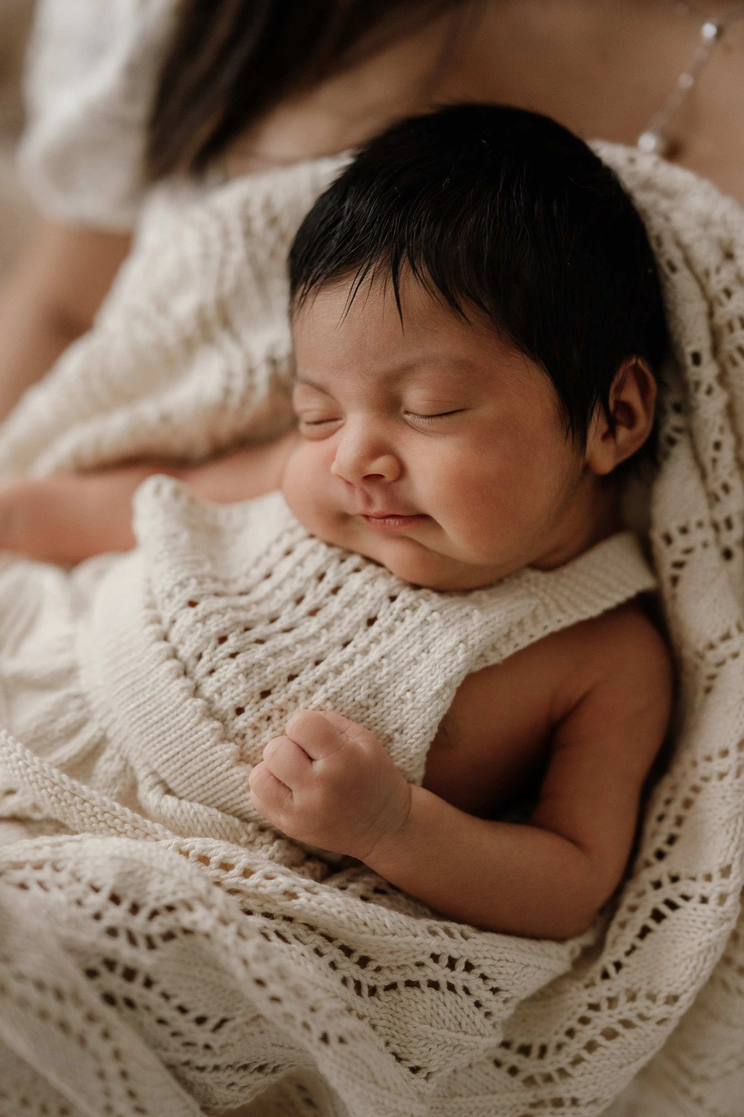 Close-up of a sleeping baby with dark hair, wrapped in a cozy beige knitted blanket, held gently by an adult.