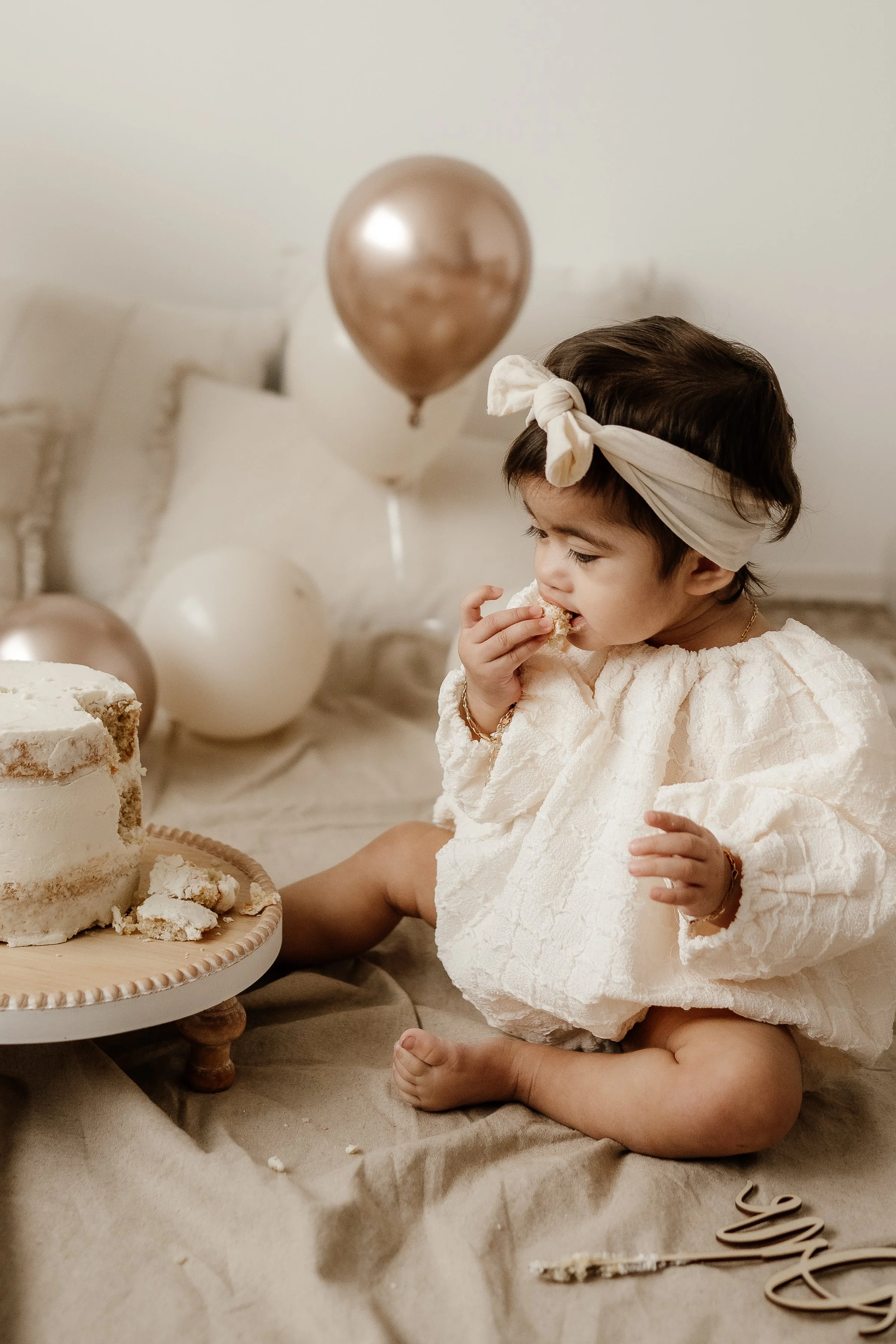 Young girl celebrating a birthday, sitting on the floor, eating a piece of cake, with balloons in the background.