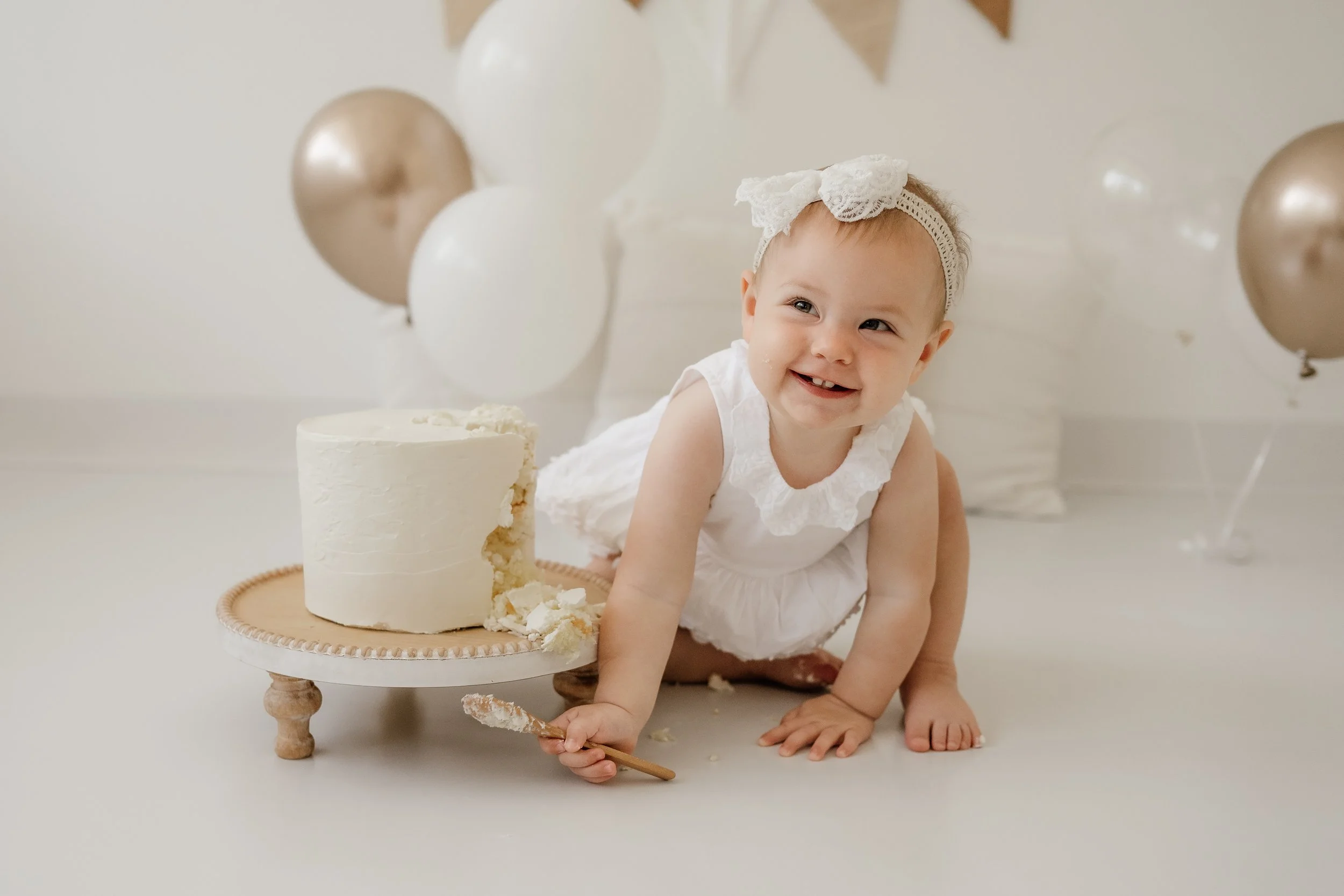 A smiling baby girl with a white lace headband in a white dress, crawling on the floor with a partially eaten white cake on a cake stand, surrounded by white and gold balloons, in a celebrations setting.