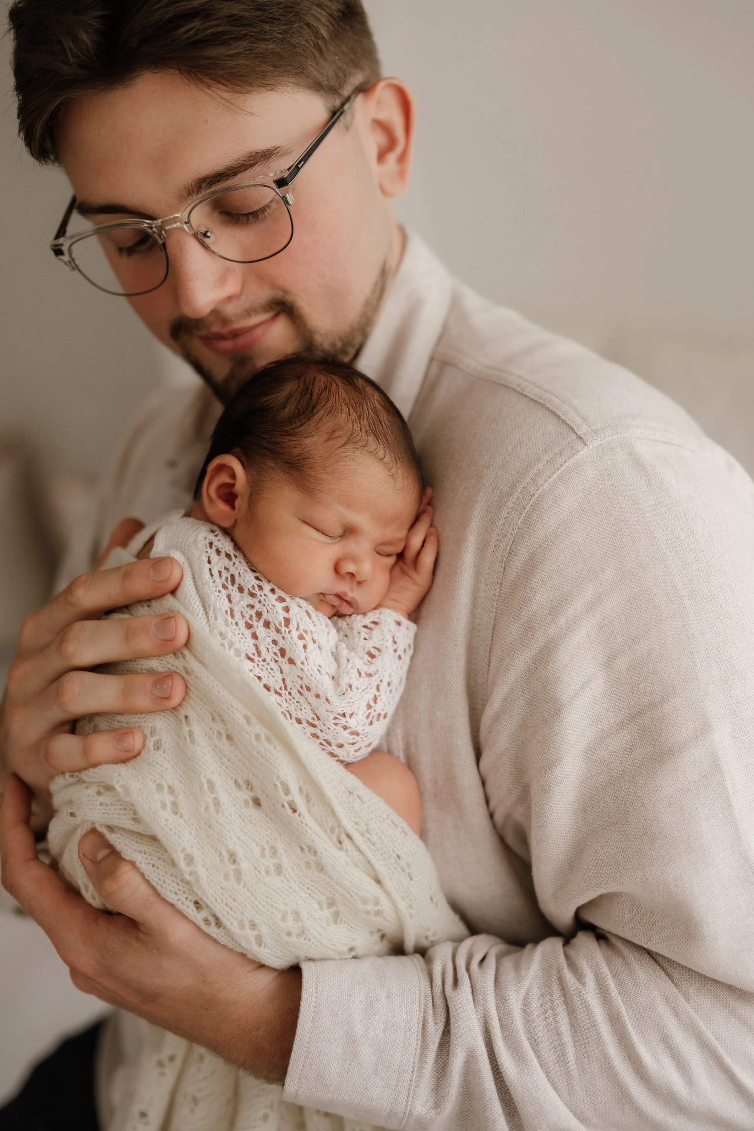 A man holding a sleeping baby wrapped in a cream-colored lace blanket, with the baby's face resting on his hand, in a warm, intimate moment.