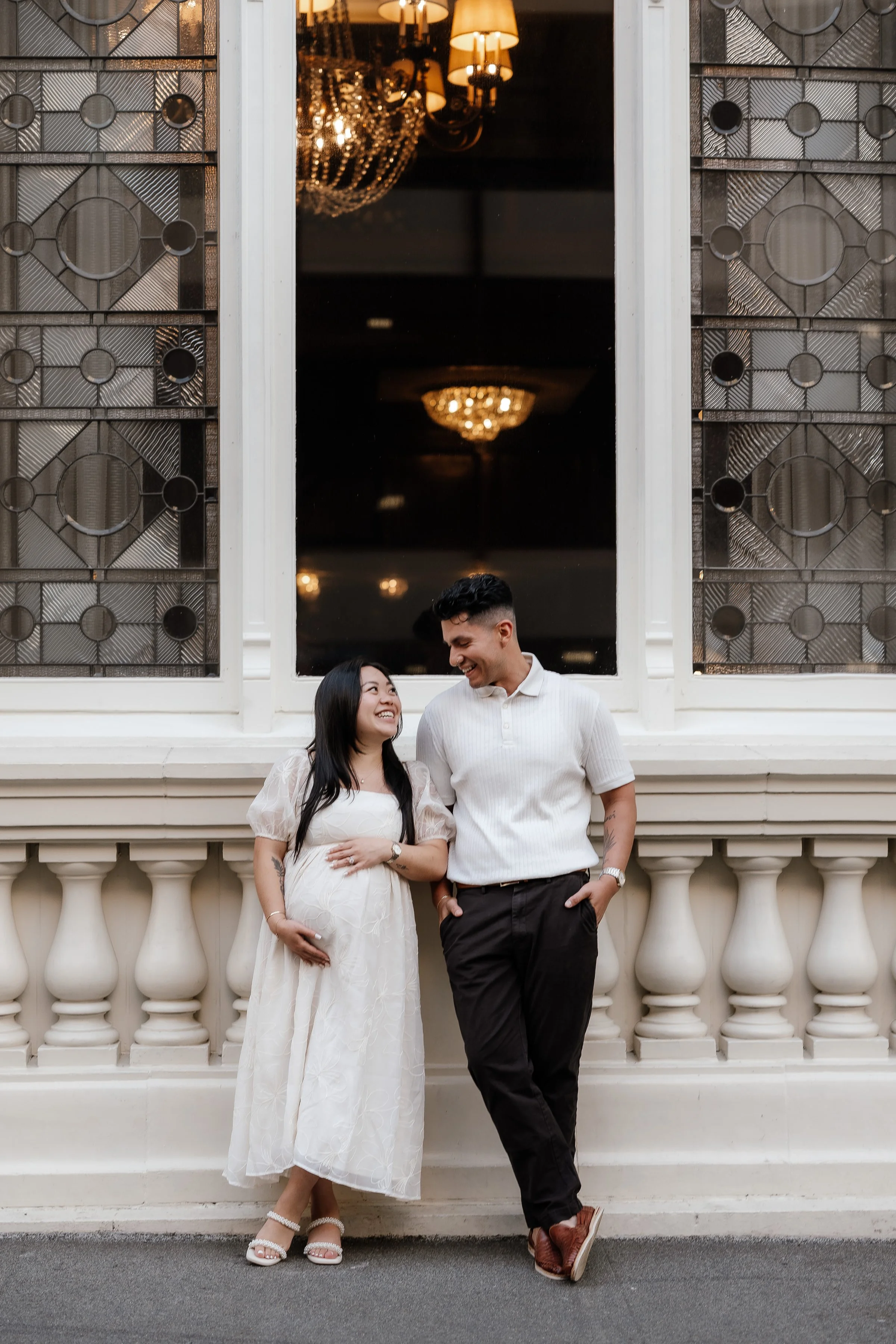 A couple standing in front of a large window with stained glass design, smiling and looking at each other. The woman is pregnant, wearing a white dress, and the man is wearing a white shirt and dark pants. Inside, elegant chandeliers are visible thro