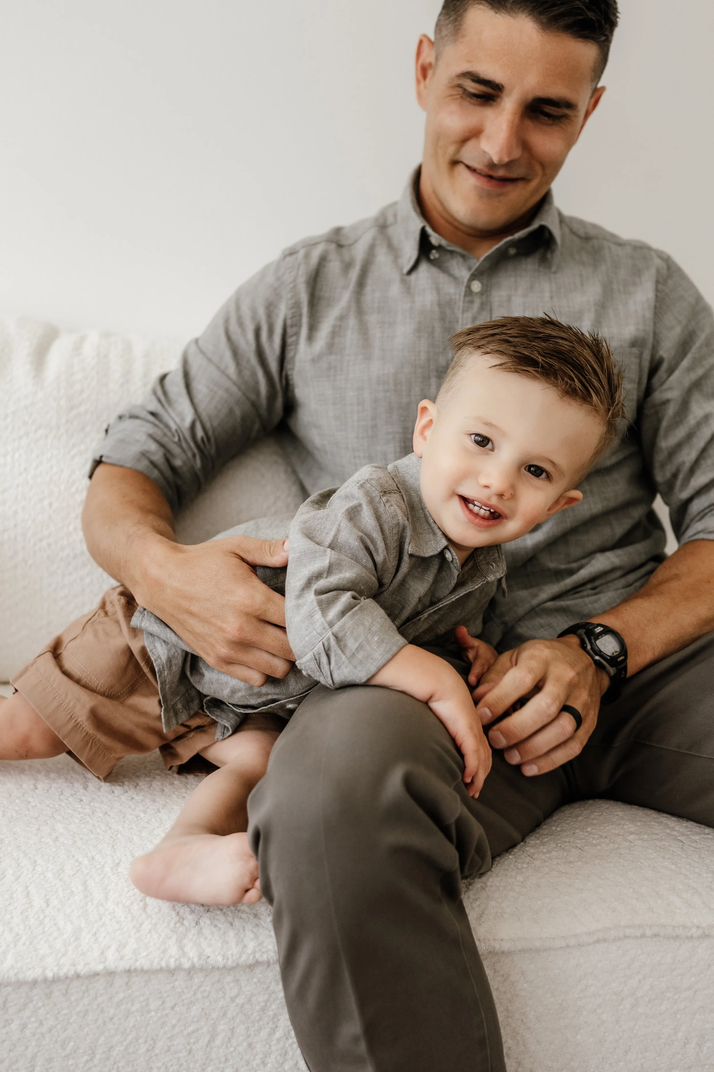 A father and young son sitting on a white couch, smiling and playing together in a cozy, well-lit room.