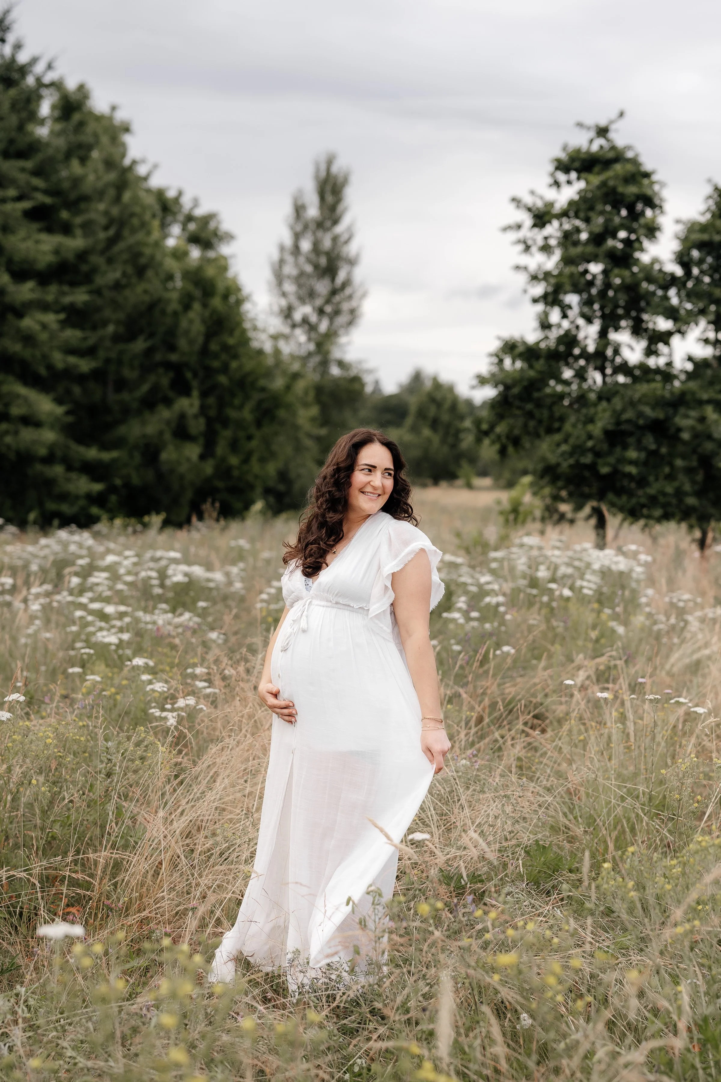 Pregnant woman in a white dress standing in a field of tall grass and wildflowers, smiling and looking to the side, with trees in the background under a cloudy sky.