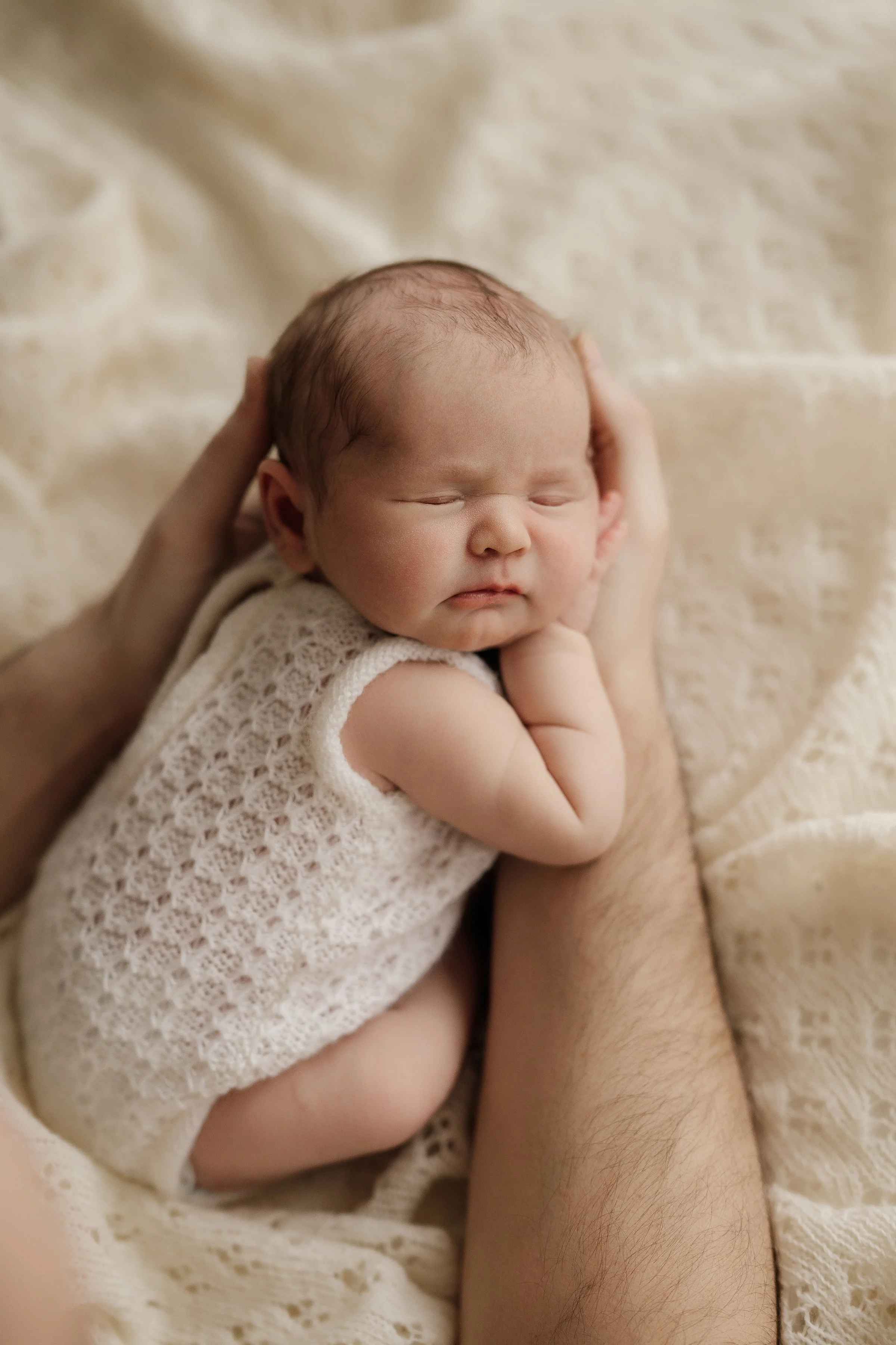 A newborn baby with closed eyes, lying on beige textured blankets, wearing a cream knitted romper, with an adult's hand supporting the baby’s head and arm.