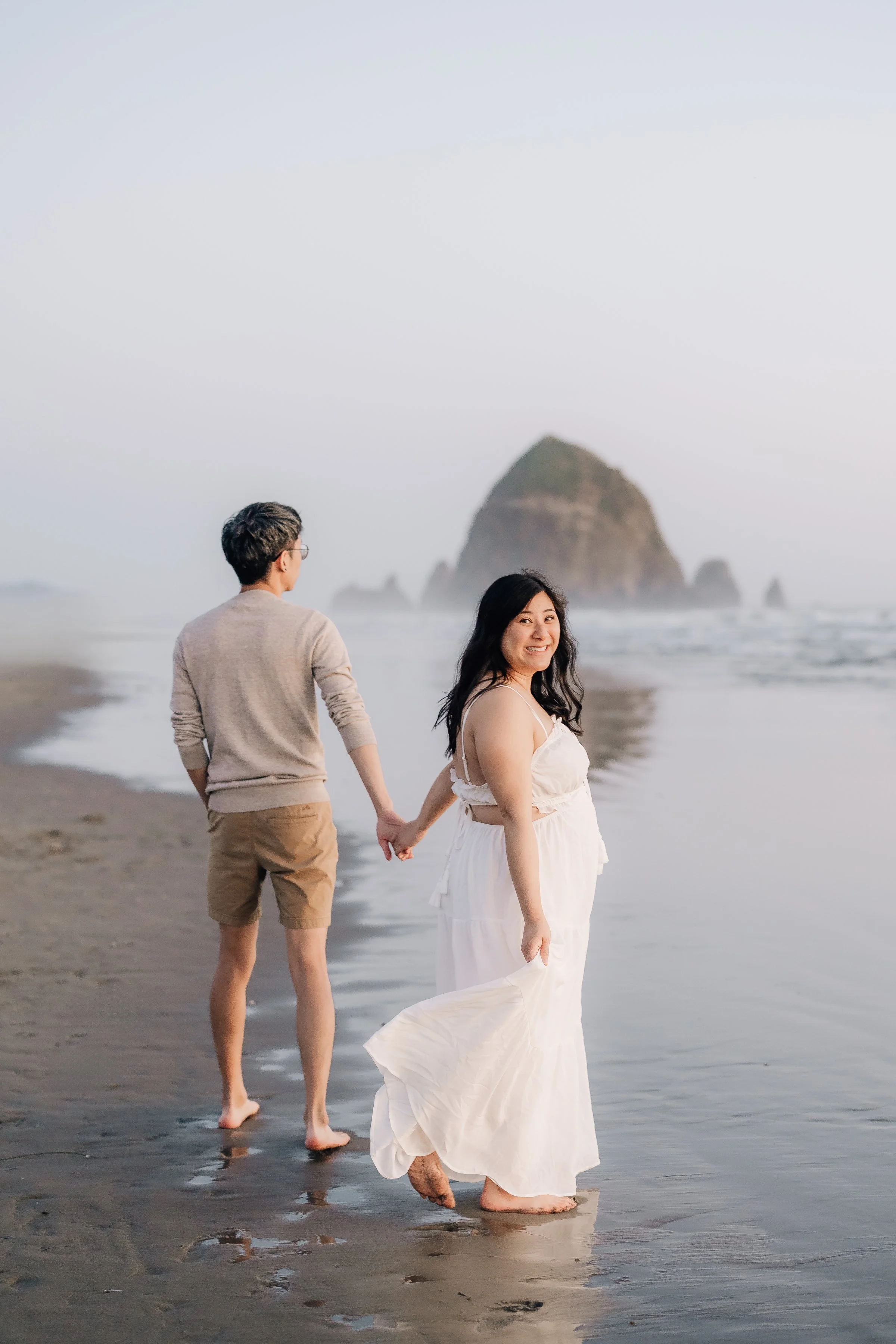 A couple walking on a beach, holding hands. The woman in a white dress is smiling at the camera, while the man is facing away. There is a large rock formation in the background.