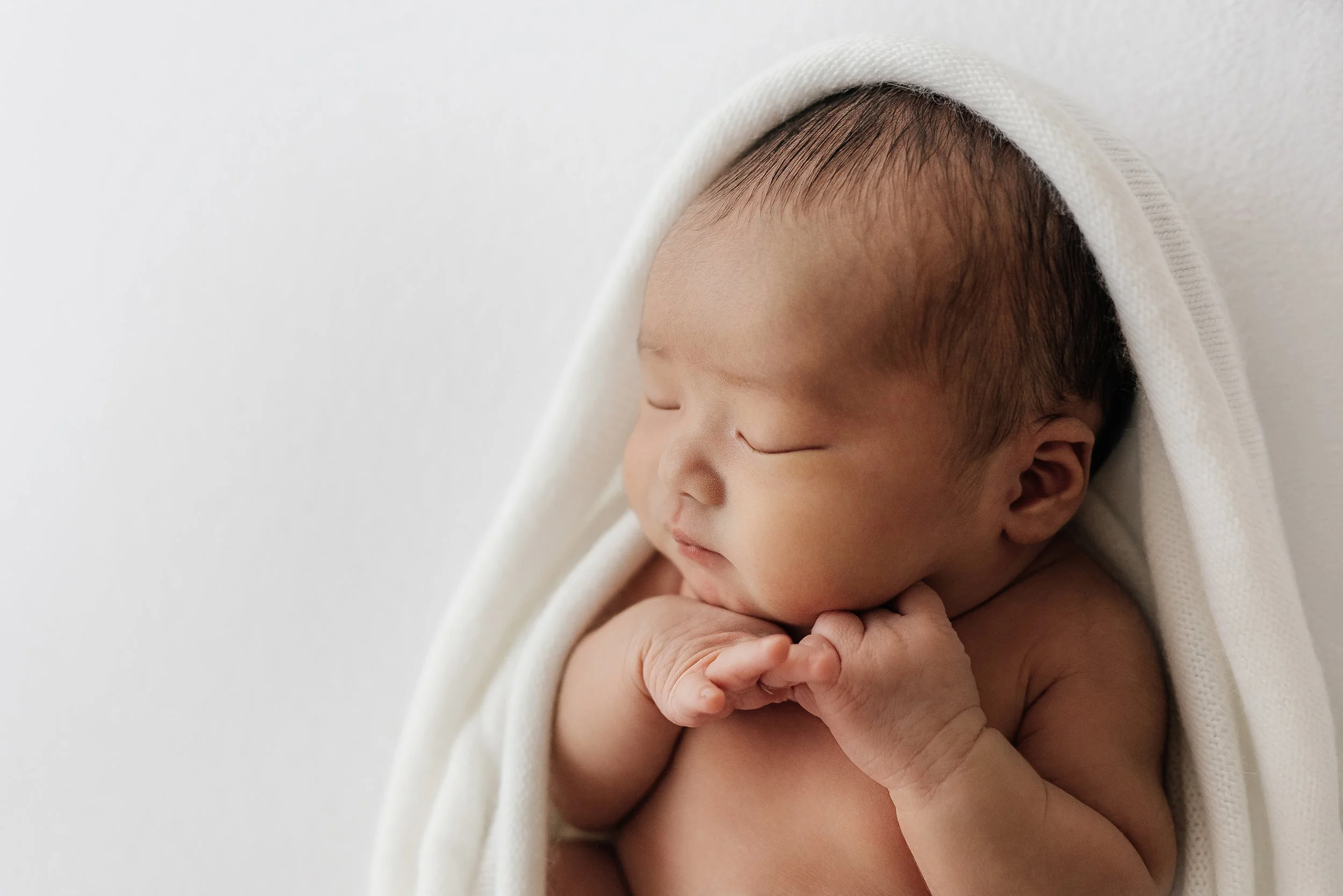 Newborn baby peacefully swaddled in white during a natural light studio session.