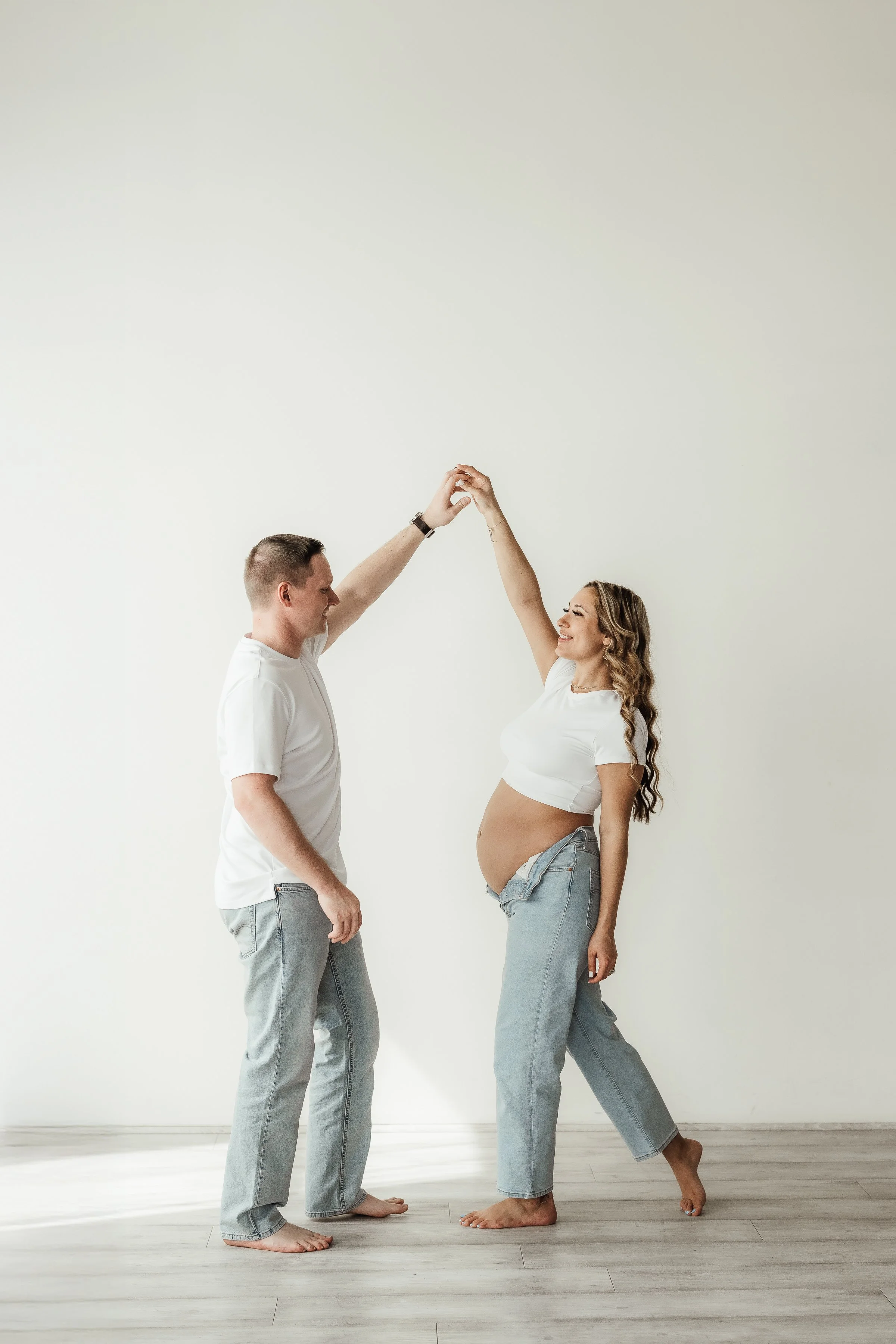 A happy pregnant woman and her partner holding hands, smiling, and dancing together in a minimalist room with light-colored hardwood floors and plain white walls.
